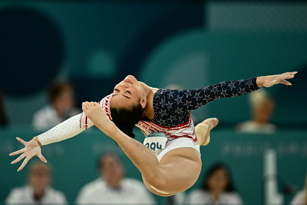 A gymnast performs an intricate routine mid-air, wearing a star-patterned leotard. The audience in the blurred background watches intently
