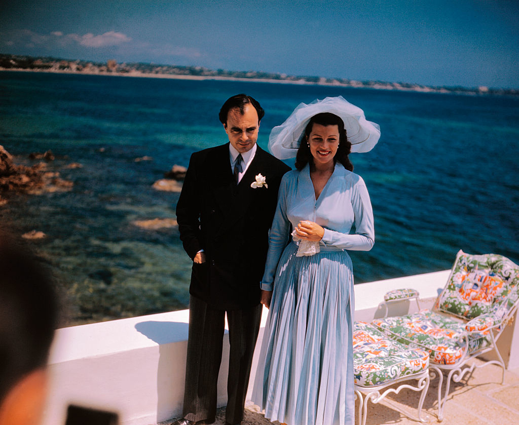 Aly and Rita standing together by the ocean; she wears a wide-brimmed hat and a long dress, and he is in a dark suit with a boutonnière
