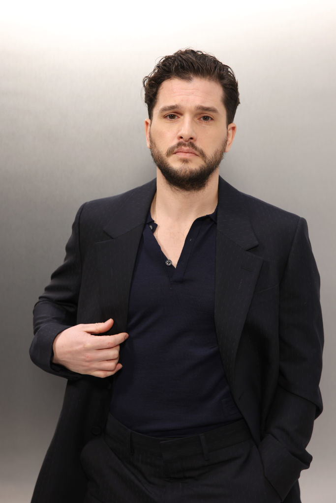 Kit Harington posing in a dark suit over a buttoned shirt, hand in pocket, against a plain background