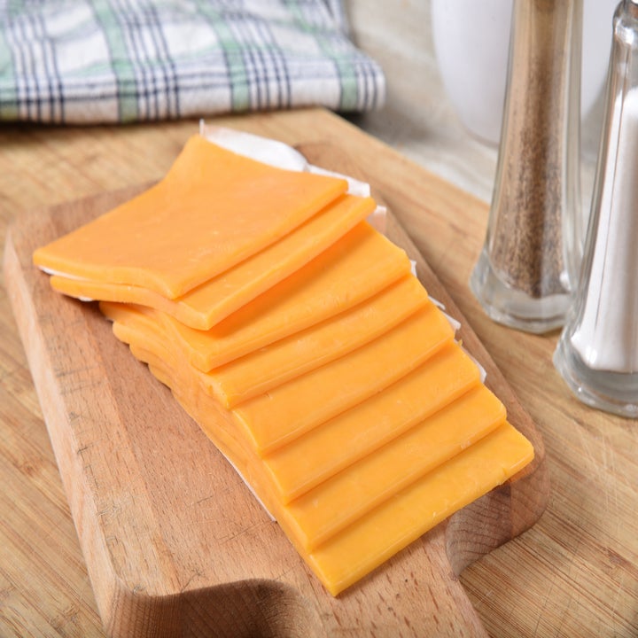 Slices of cheddar cheese are laid out on a wooden cutting board with a cloth and salt and pepper shakers in the background