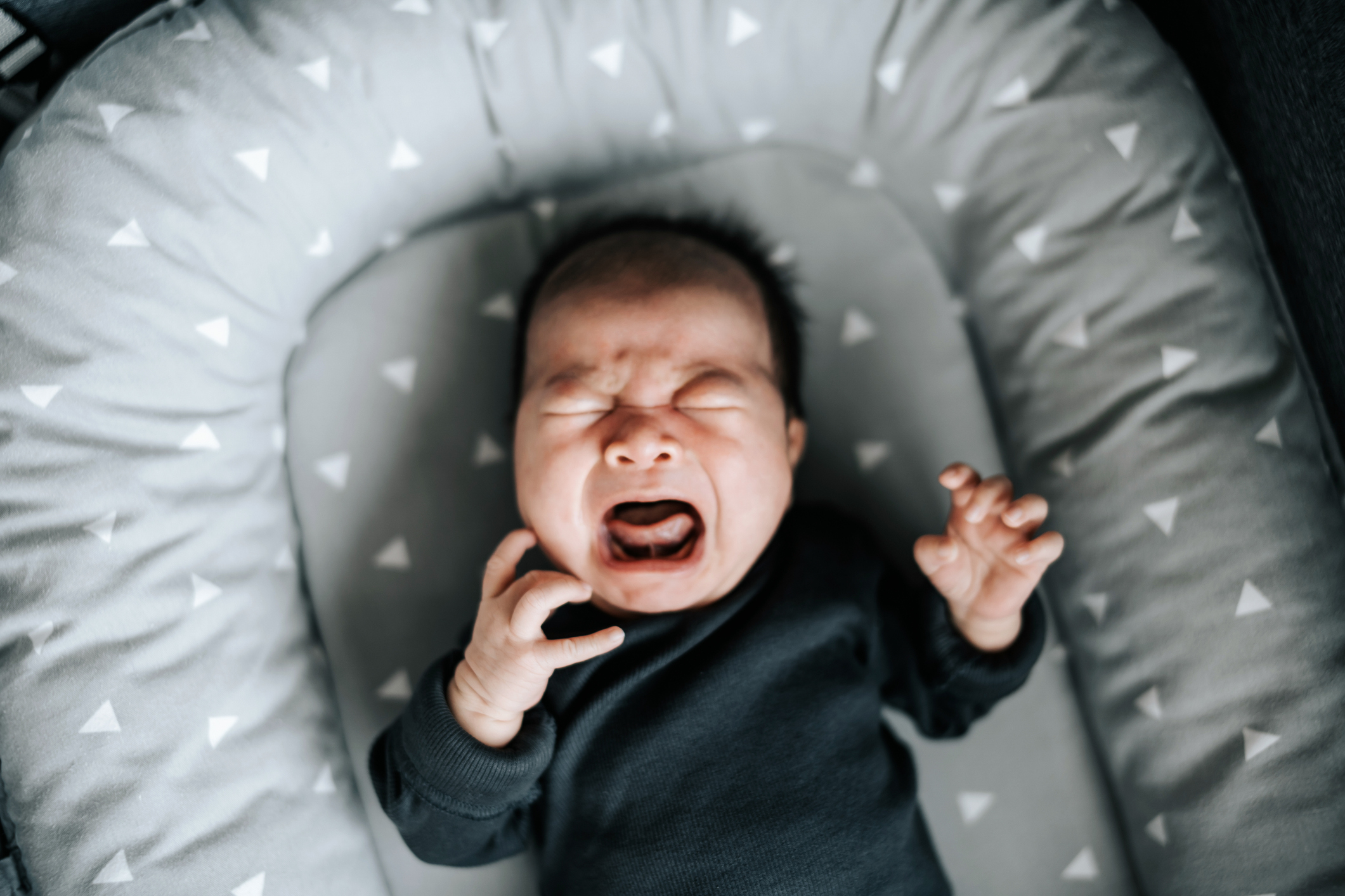 A crying baby lying in a cushioned crib. The baby wears a dark outfit and appears to be upset