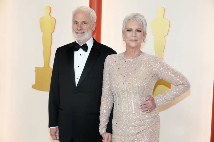 Jamie Lee Curtis in a sparkling gown poses with Christopher Guest in a tuxedo at an event with Oscar statues in the background