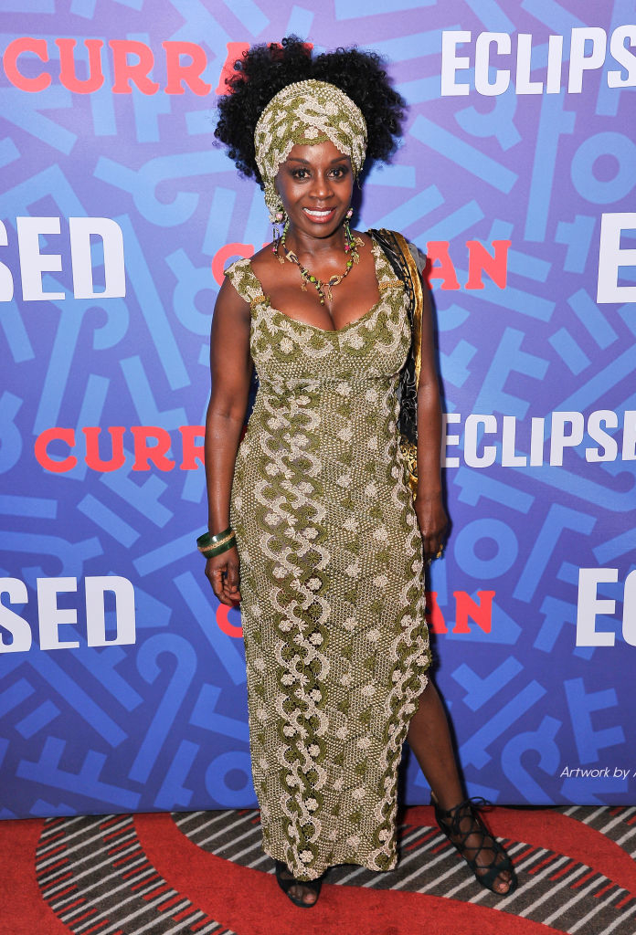 Akosua on red carpet, wearing a patterned sleeveless dress with a matching headwrap, accessorized with jewelry and heels