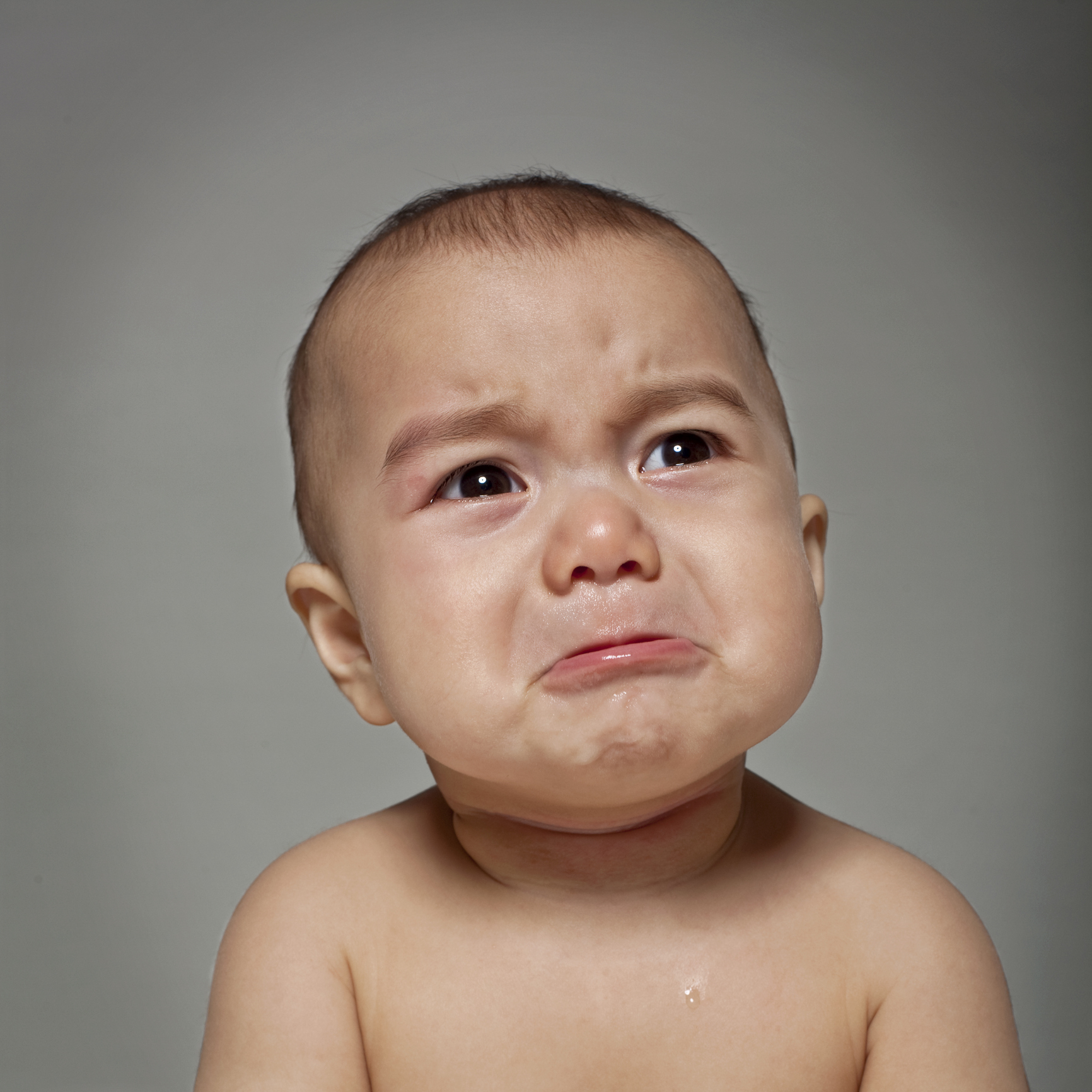A close-up of a baby crying with a distressed expression and tears on their face. The baby is shirtless with a plain background