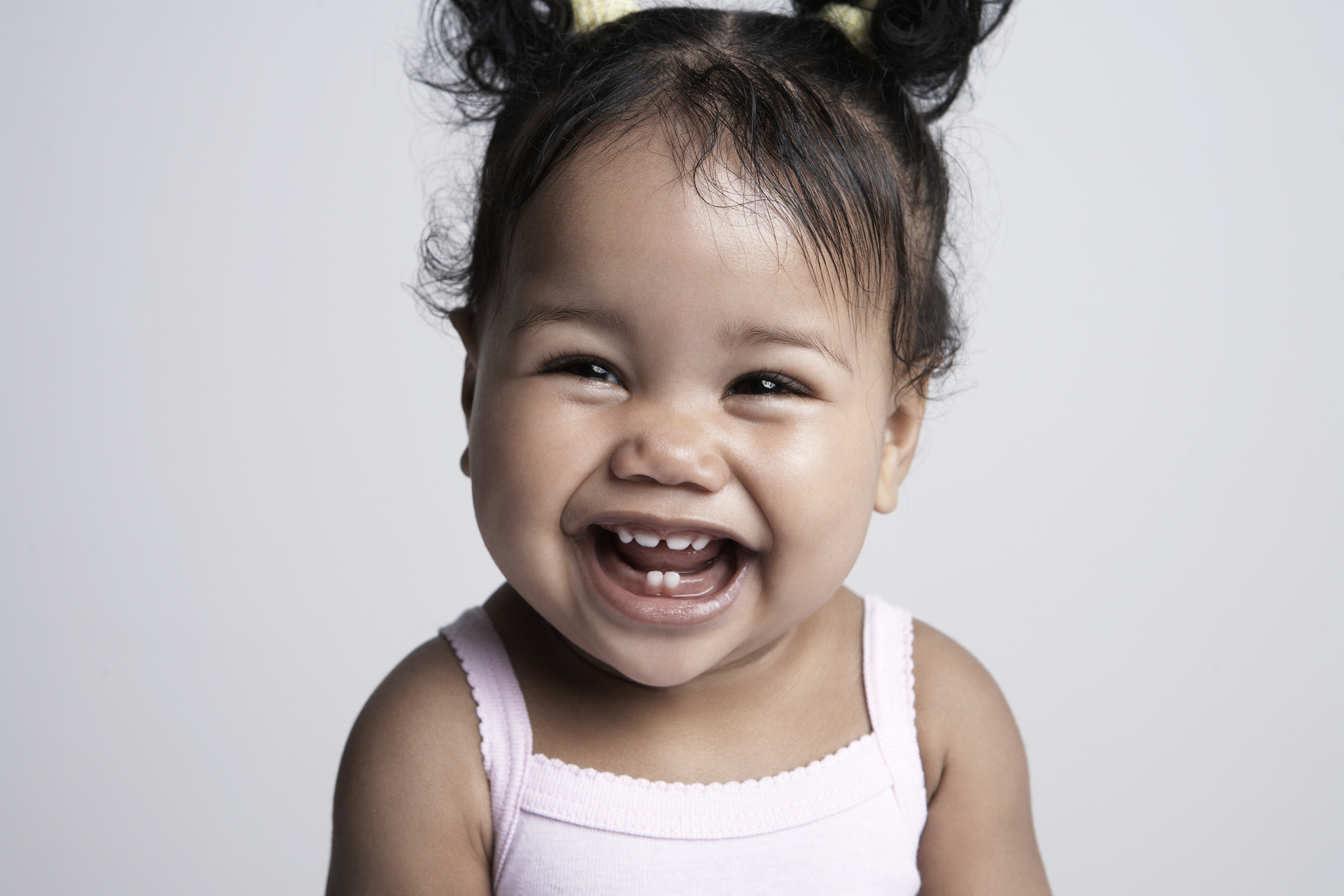 A baby with two pigtails in a sleeveless top smiles joyfully at the camera