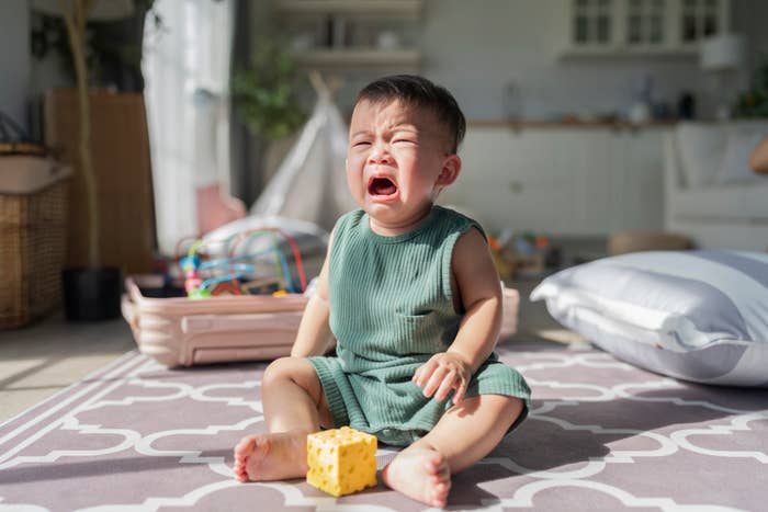 A toddler sits on a play mat, crying with an upset expression. A toy block is in their lap. Background includes toys and household items