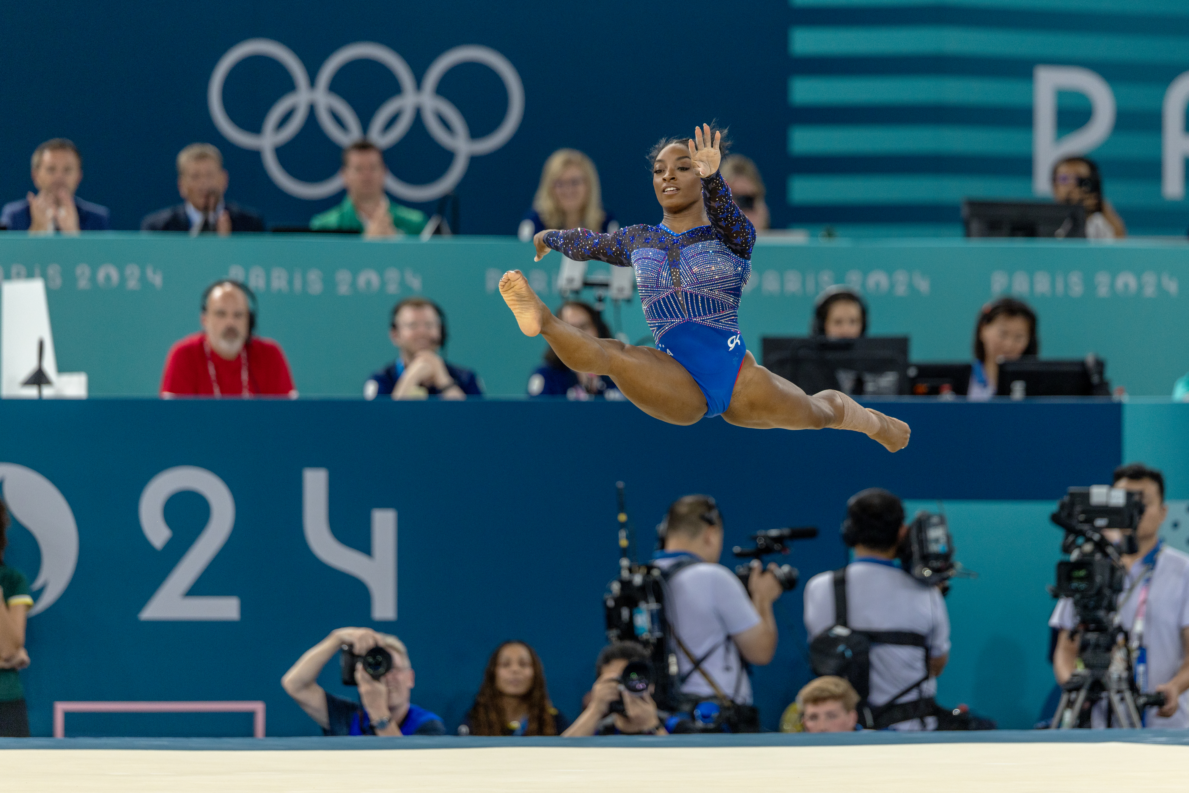 Simone Biles performing a jump during a gymnastics routine at the Paris 2024 Olympics, with photographers and judges visible in the background
