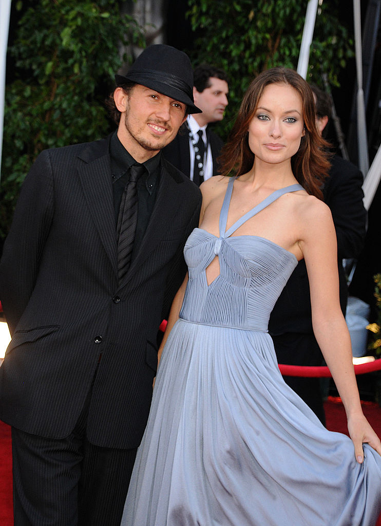 Tao and Olivia pose on a red carpet. Olivia wears an elegant, sleeveless gown with a cutout design, while Tao is in a dark suit with a fedora