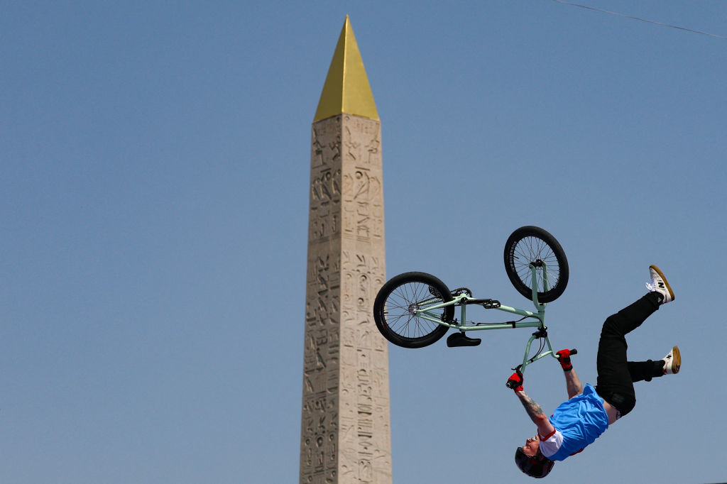 A person on a BMX performs a mid-air trick in front of a tall obelisk with hieroglyphics