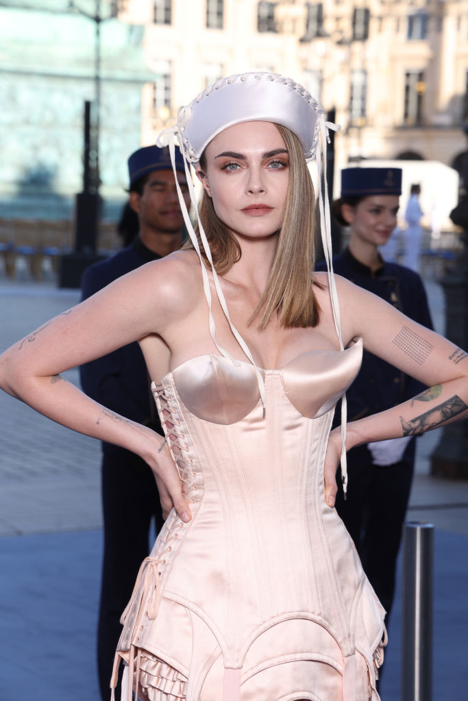 Cara Delevingne poses in a satin corset dress with detailed stitching and a matching hat, hands on hips, attending an outdoor event