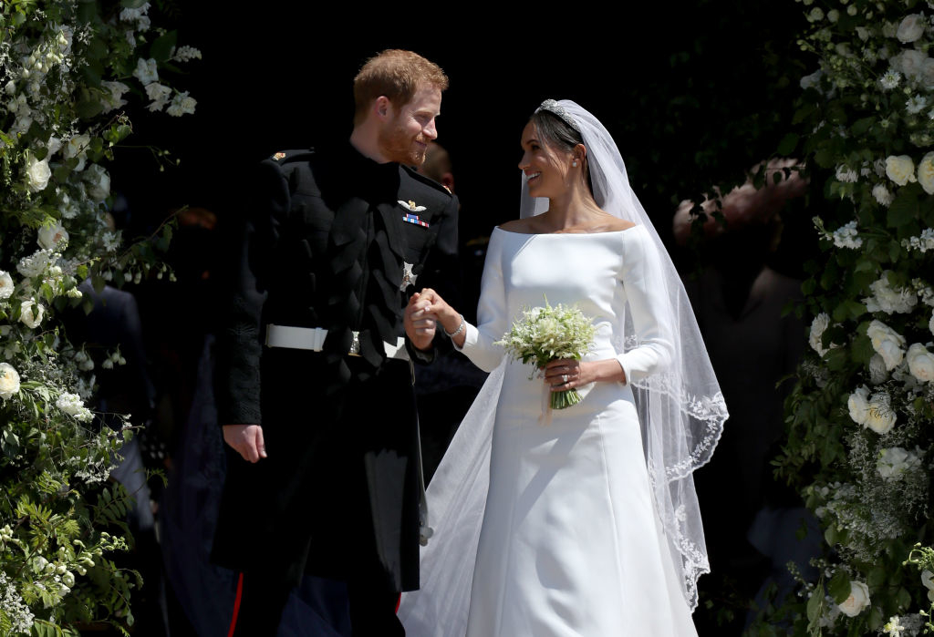 Prince Harry and Meghan Markle smiling and holding hands, standing under a floral arch on their wedding day. Harry in military uniform, Meghan in an elegant wedding dress