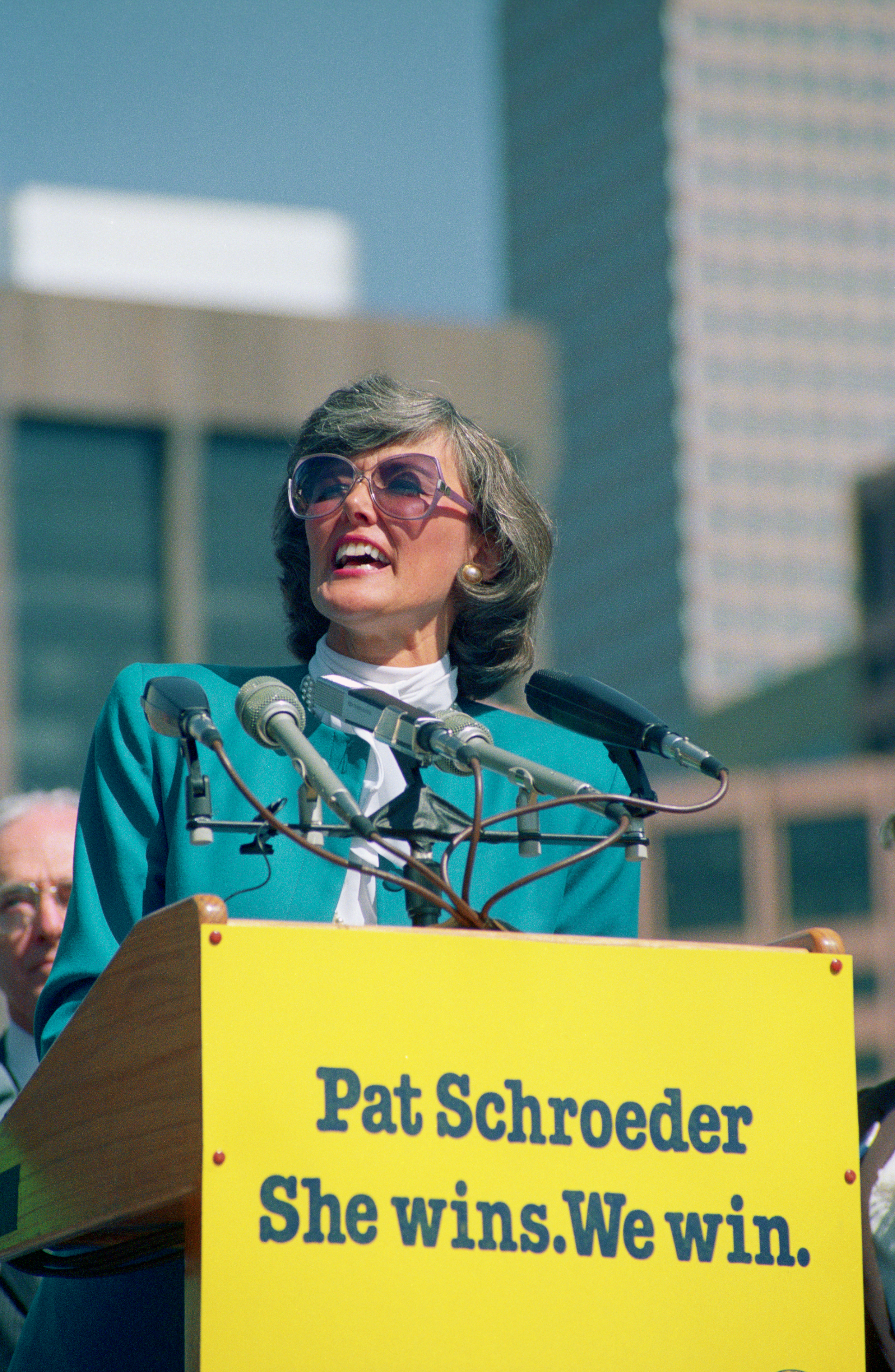 Pat Schroeder stands at a podium with microphones, delivering a speech. The podium has a sign that reads, "Pat Schroeder She wins. We win."