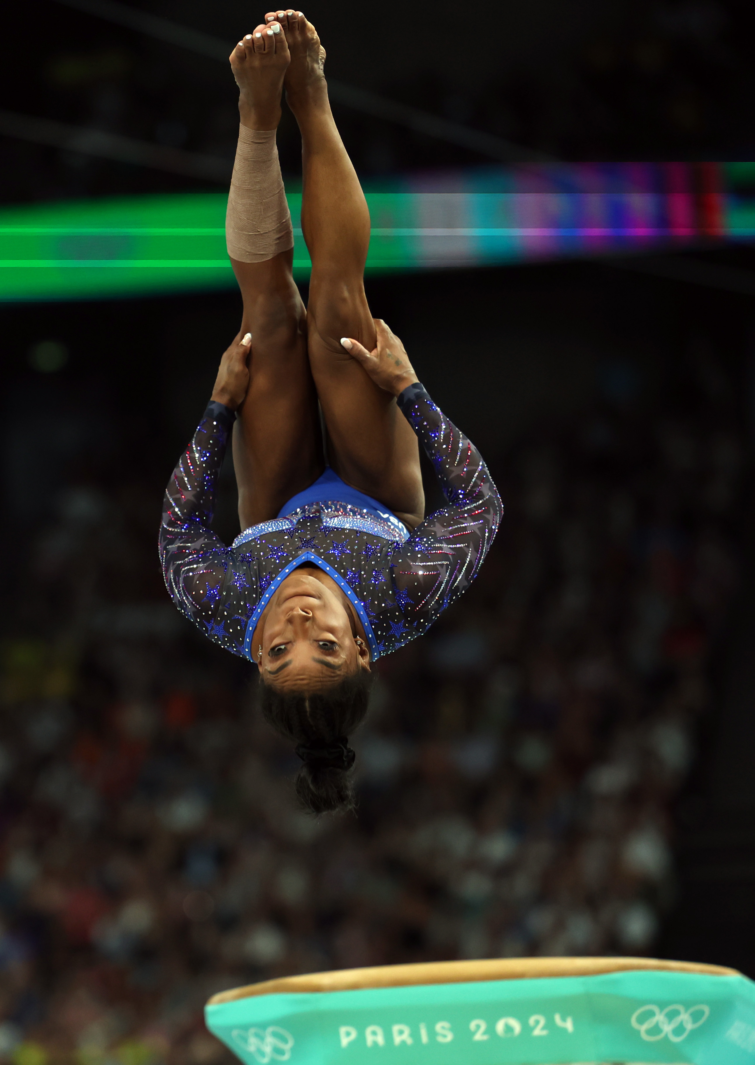 Simone Biles performing a high-flying gymnastics routine, upside down with legs bent and wrapped in a star-studded outfit at the Paris 2024 Olympics