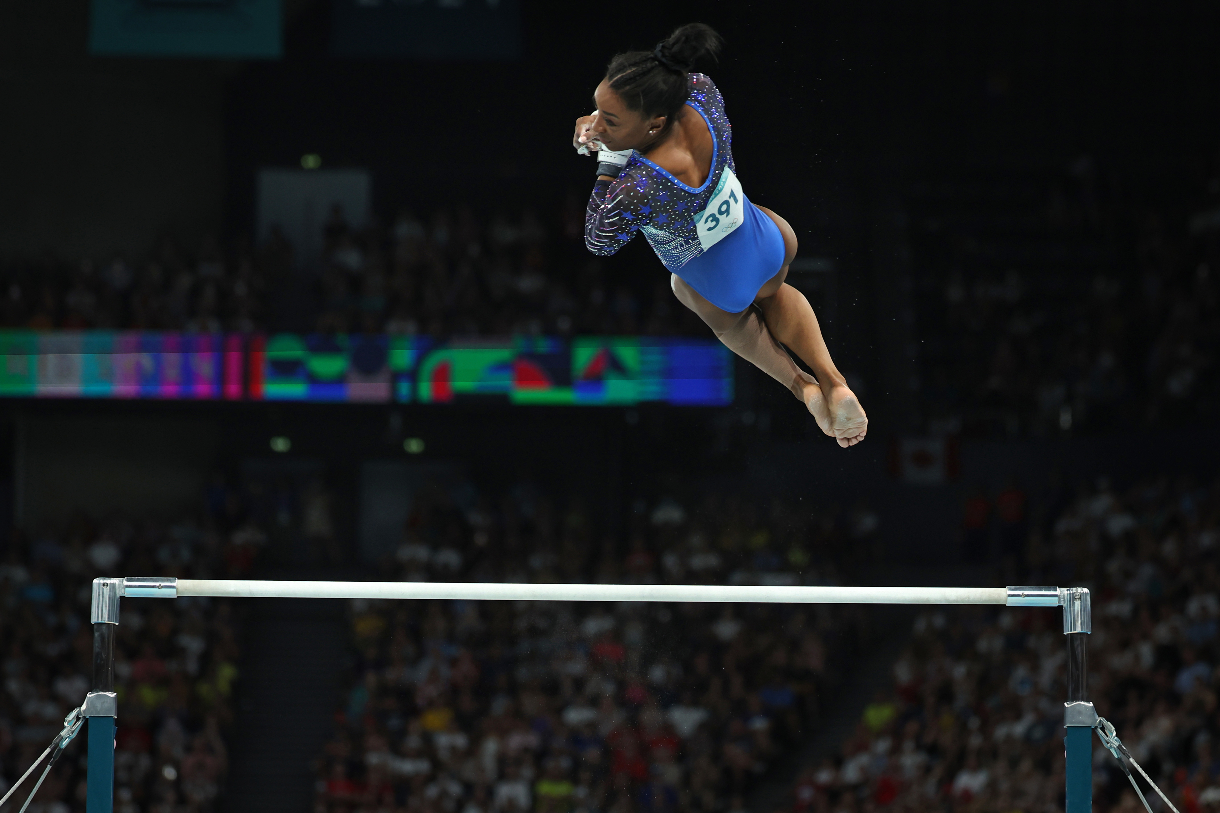 Simone Biles mid-air during a routine on the uneven bars during a gymnastics competition. She is mid-air, wearing a leotard and gripping the bars