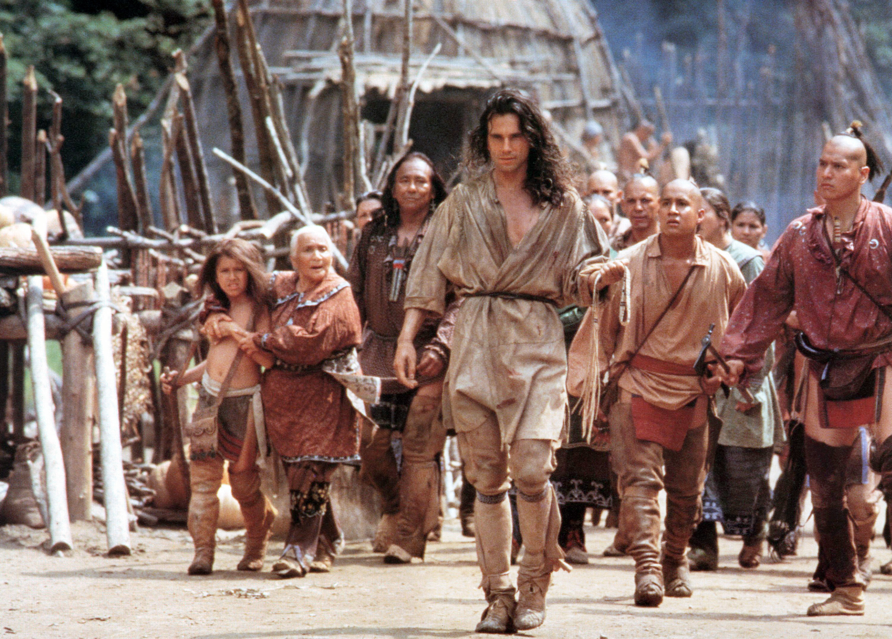 Daniel Day-Lewis, in rustic attire, leads a group of people in historical attire, staged outdoors in front of wooden huts