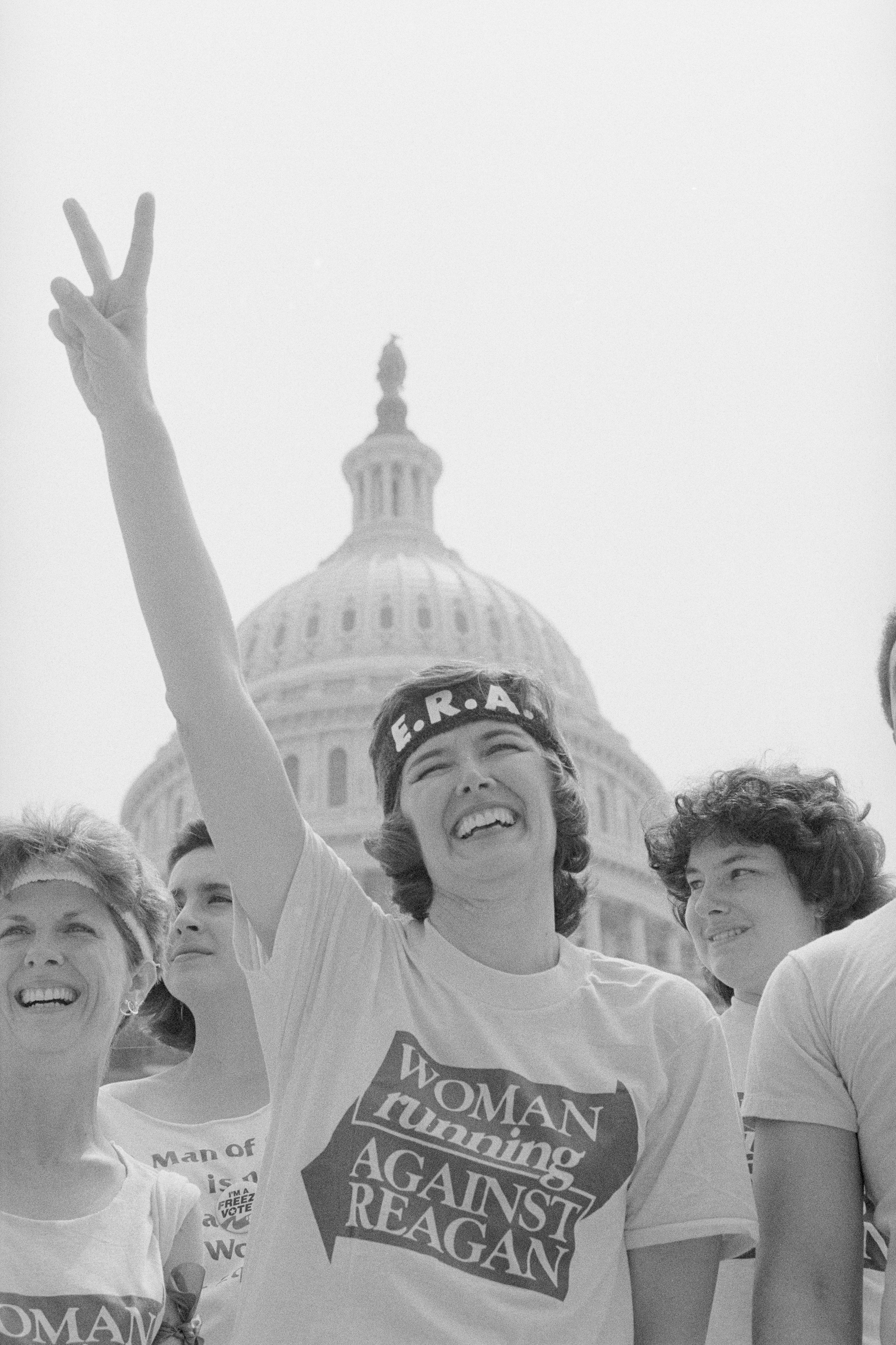 A group of women, including one wearing an ERA headband and "Woman Running Against Reagan" shirt, in front of the U.S. Capitol, raising a peace sign