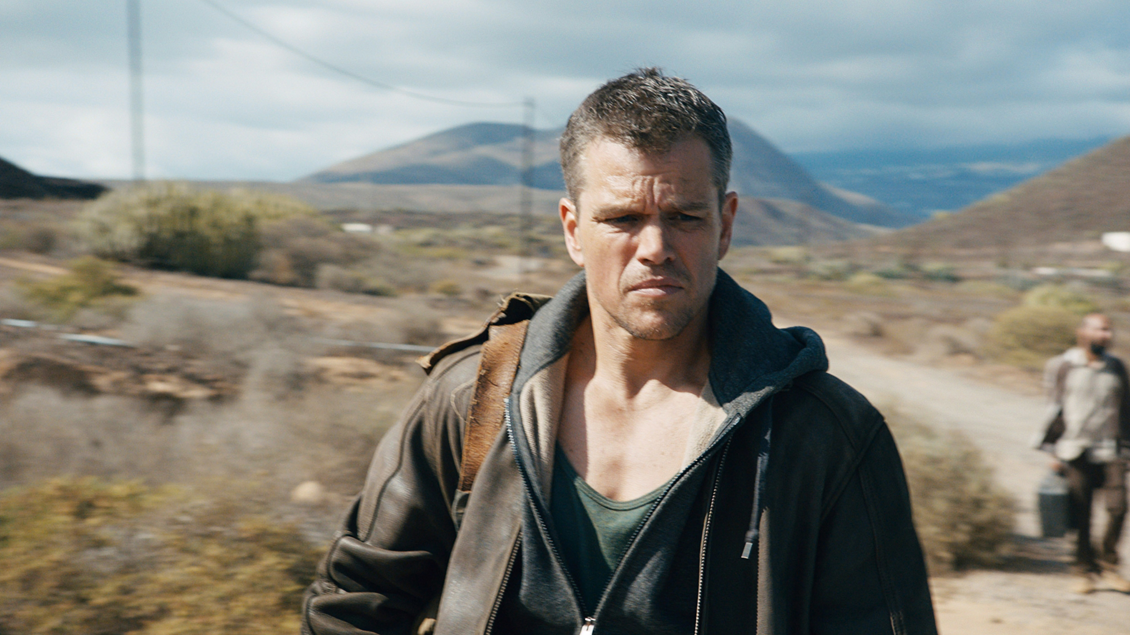 Matt Damon walks in a rugged landscape, wearing a leather jacket and a hoodie, with a serious expression