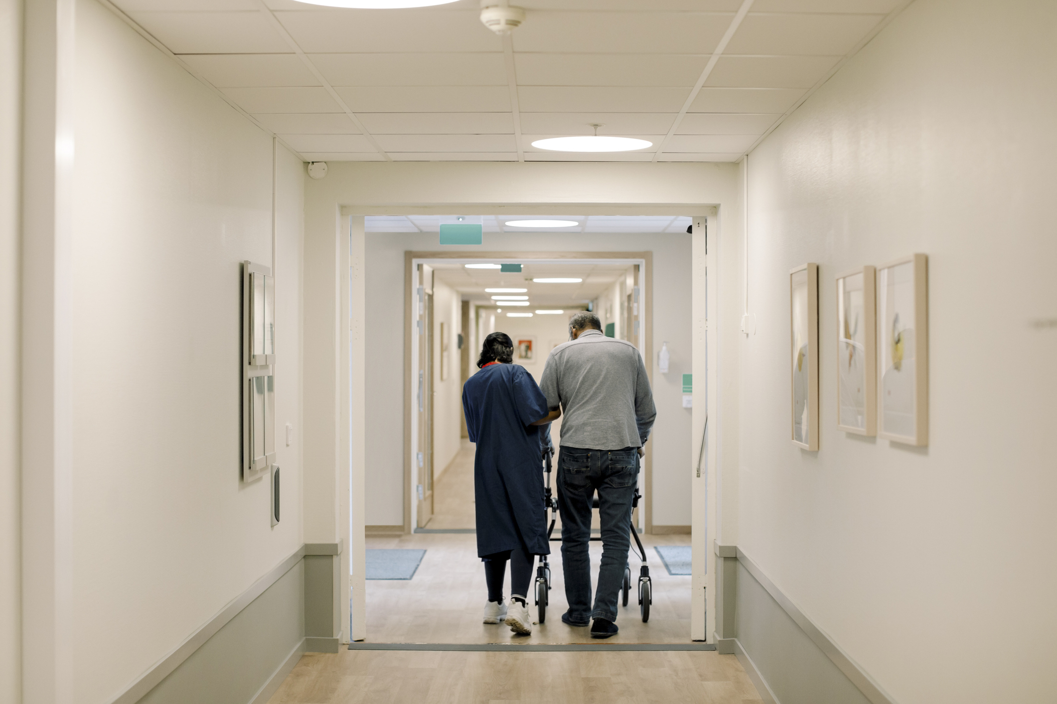 Two adults walking down a well-lit hallway in a healthcare facility. One individual assists the other using a walking aid