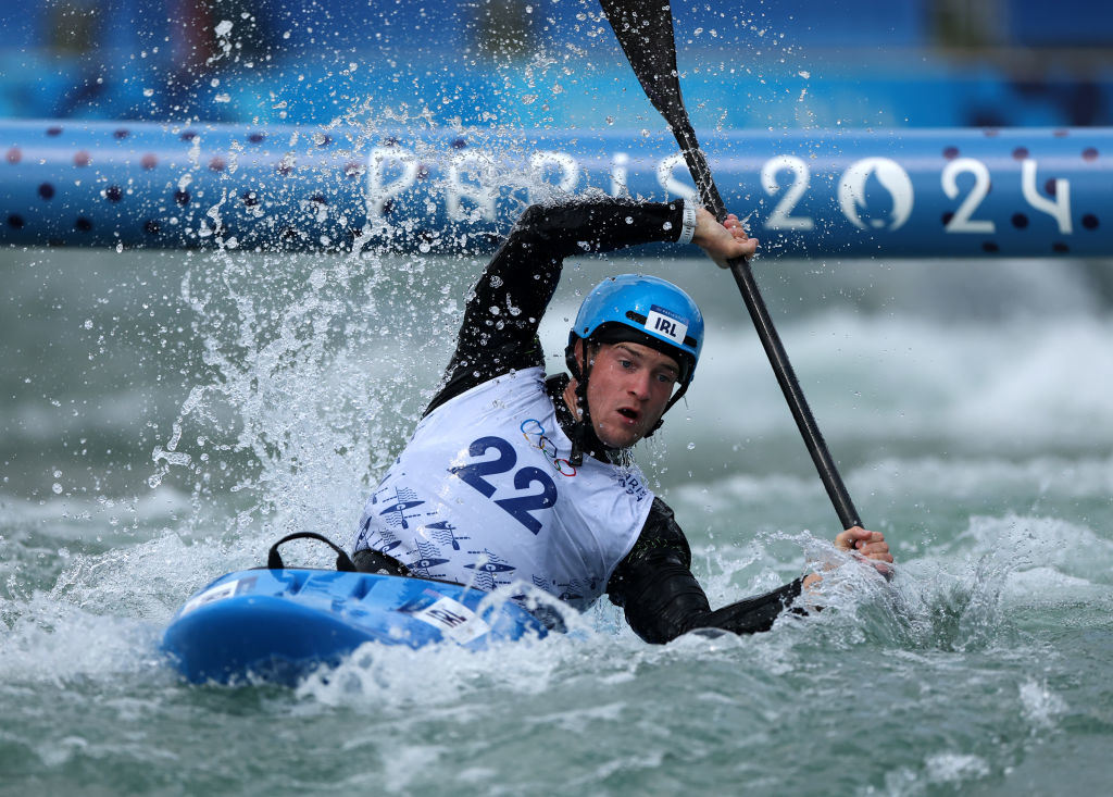 Person wearing a blue helmet and white jersey with number 22 kayaking in turbulent water, with "Paris 2024" visible in the background