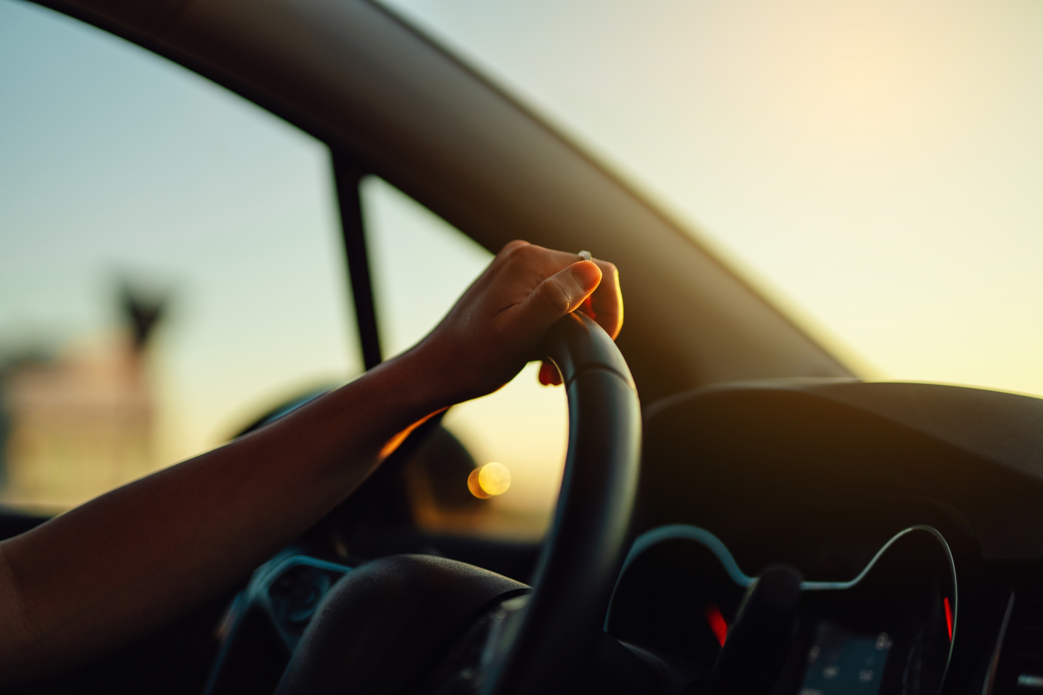 A person's hand is holding the steering wheel of a car, with the sun setting in the background. The image is serene and focuses on driving during sunset