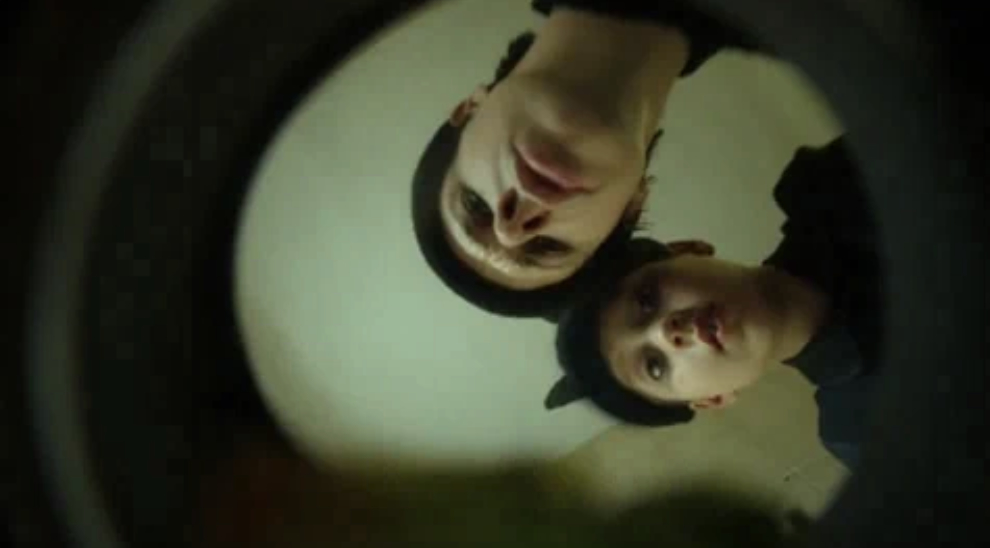 Two people are looking downwards into the camera, creating an unusual, slightly distorted perspective. Their expressions appear serious or curious