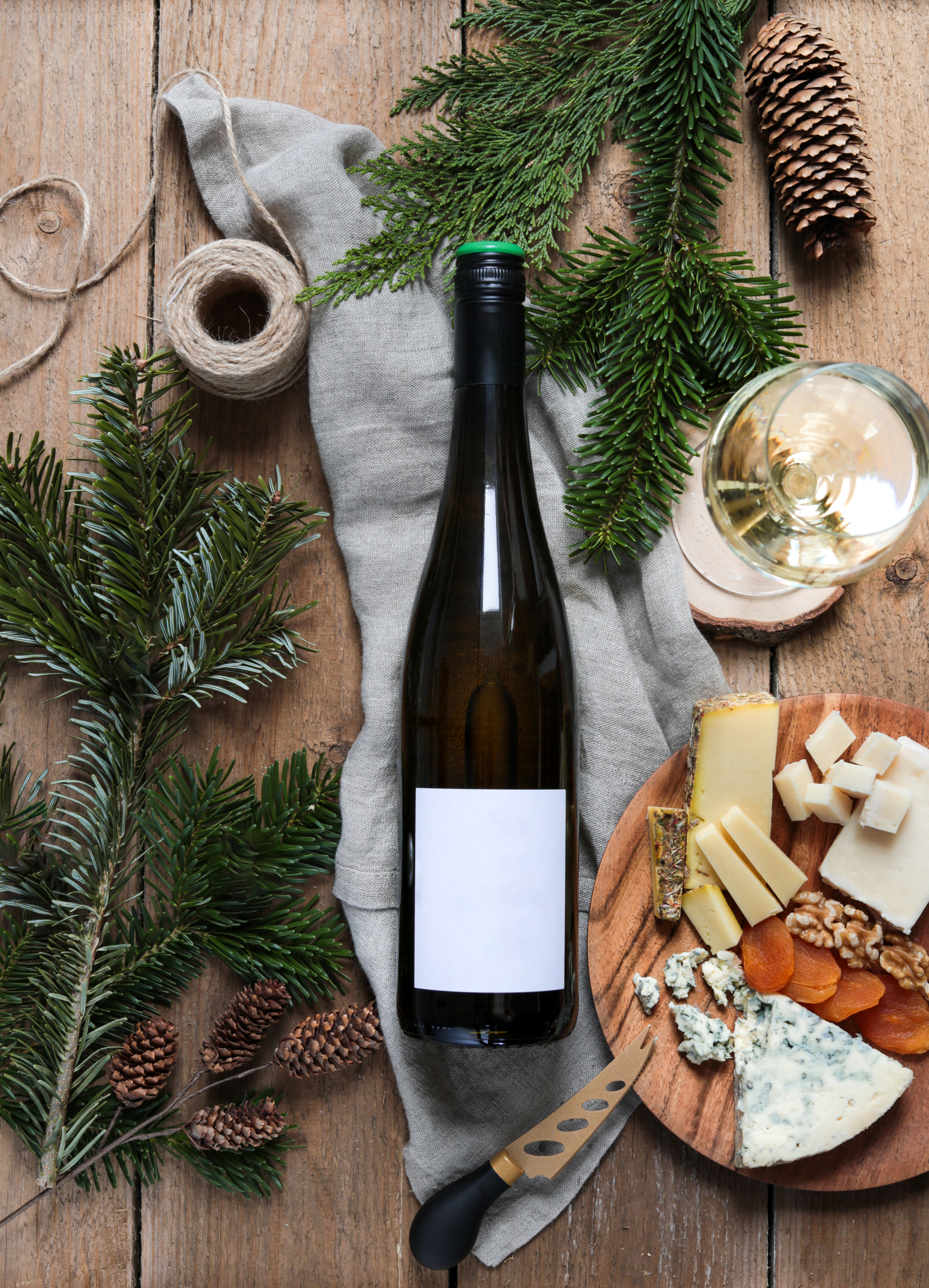 A bottle of wine on a rustic wooden table with cheese, nuts, dried apricots, a knife, a glass of wine, and sprigs of evergreen