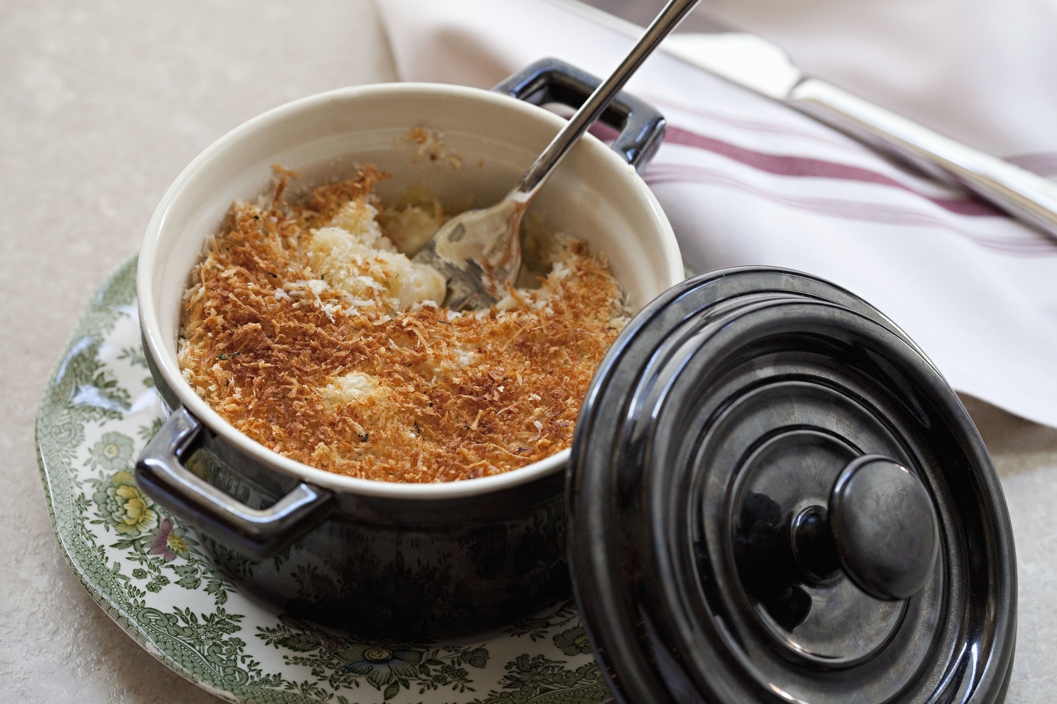 A baked dish with a golden-brown crust served in a small black pot on a floral plate, next to a white napkin with red stripes and a silver fork