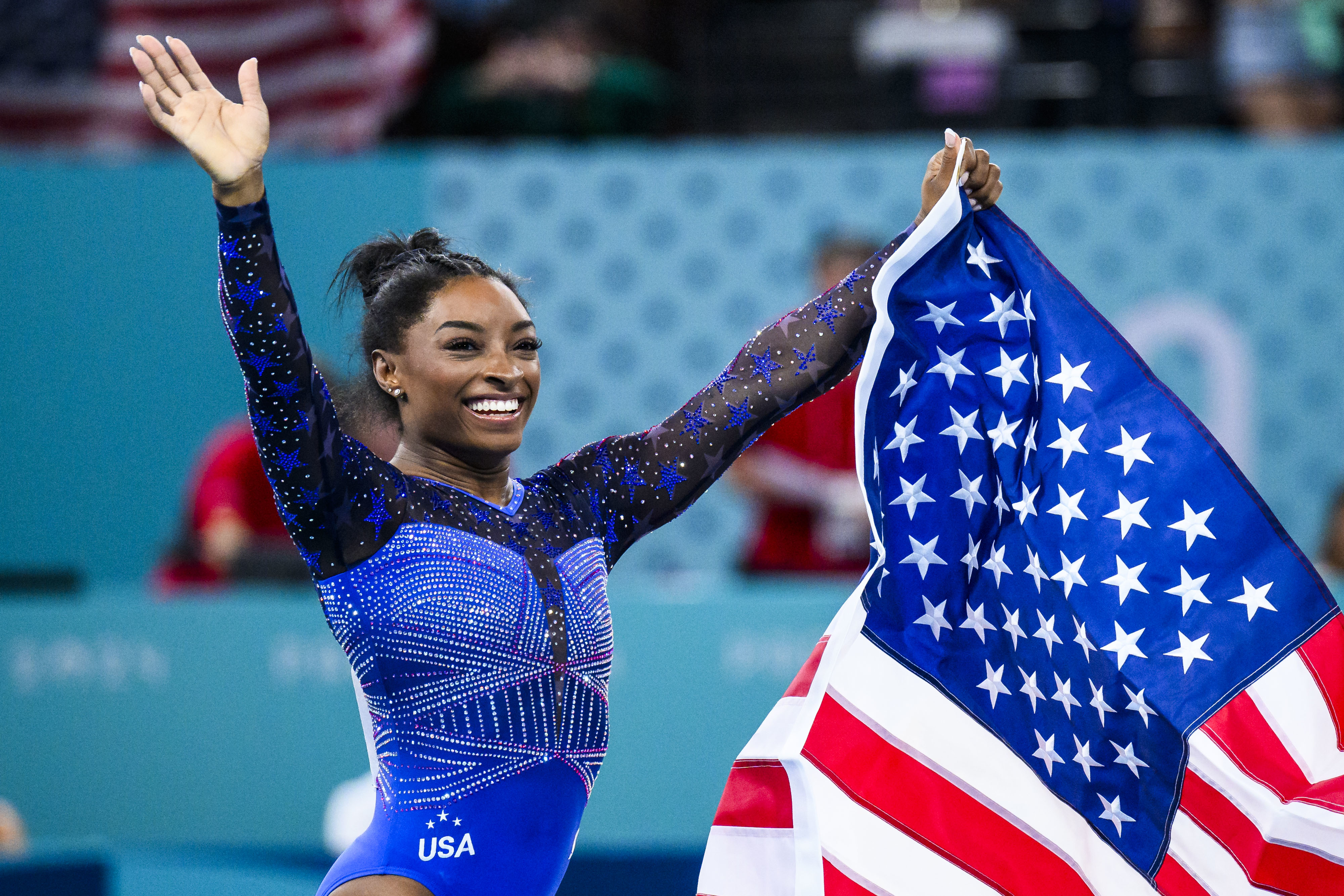Simone Biles in a sparkling leotard waves and holds an American flag, celebrating a gymnastics victory