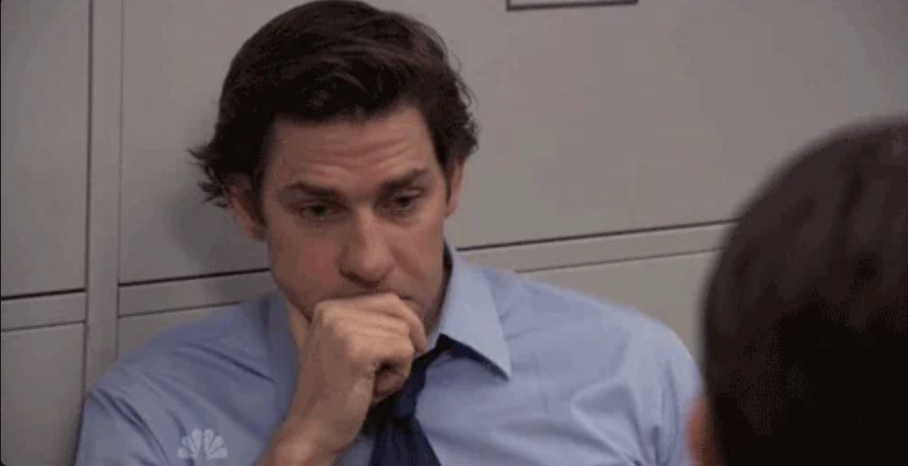 John Krasinski, wearing a business dress shirt, appears pensive with his hand resting on his chin in an office setting