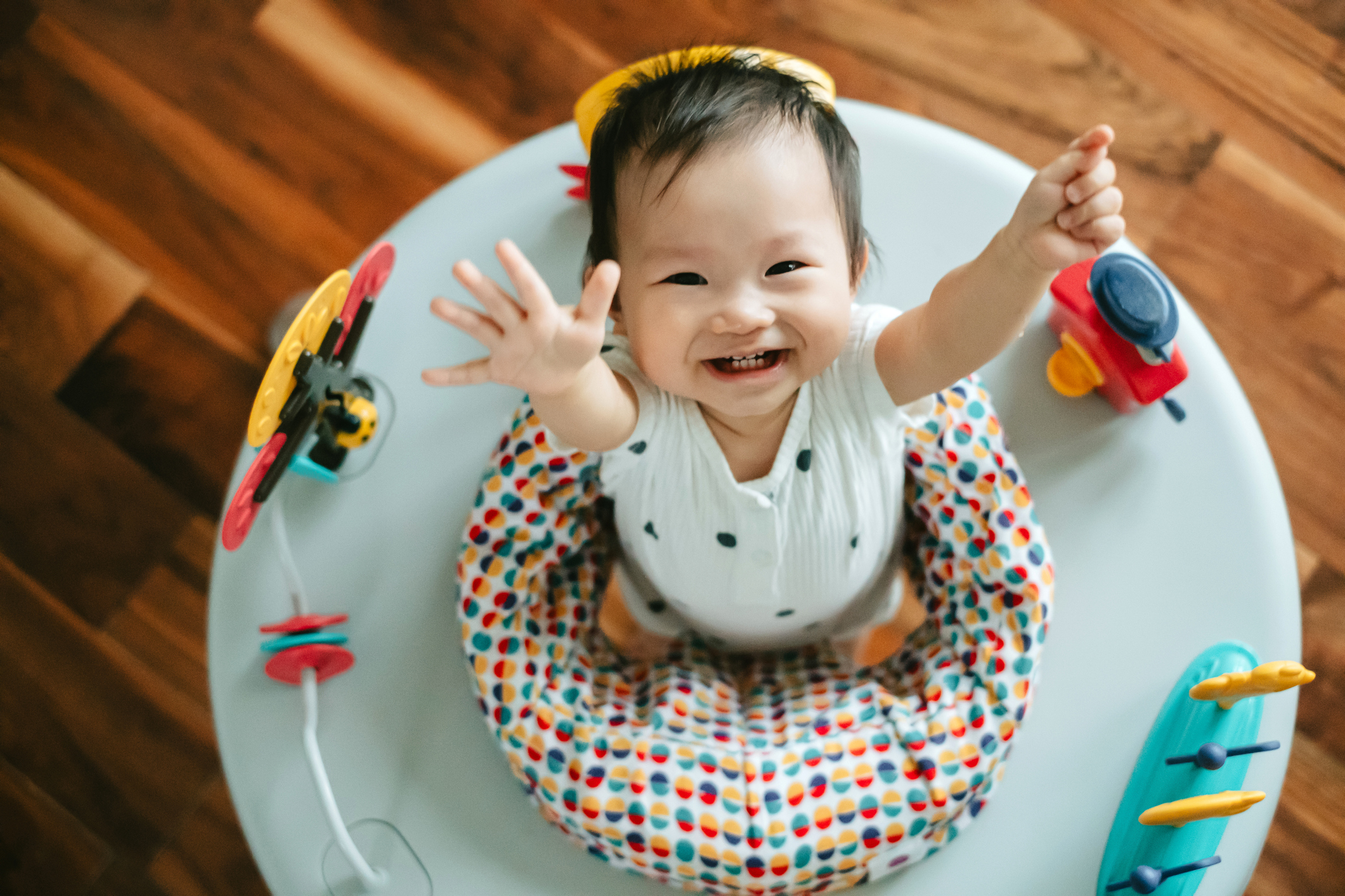 A smiling baby in a polka-dot outfit reaches up while seated in a colorful baby activity center
