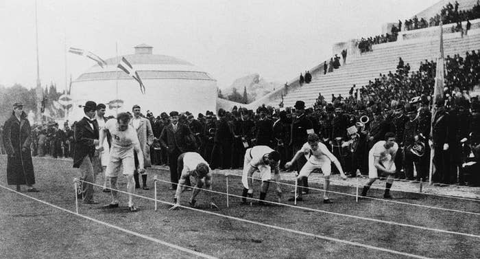 Track and field athletes crouch at the starting line in a historic race, with a large crowd of spectators watching from the stands and a building in the background