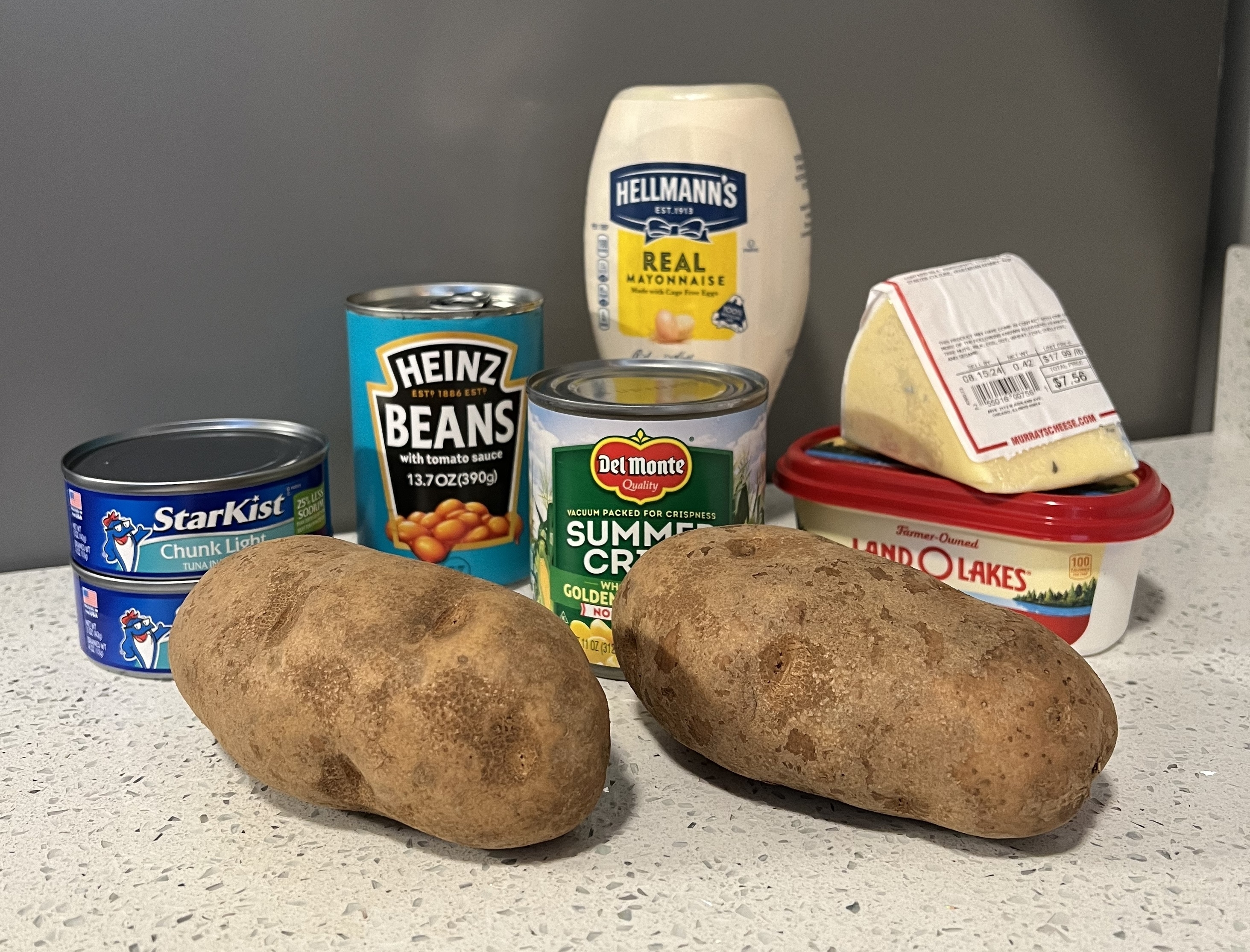 Two russet potatoes sit on a countertop surrounded by Heinz beans, Hellmann's mayonnaise, Starkist tuna, Del Monte corn, cheese, and Land O'Lakes butter