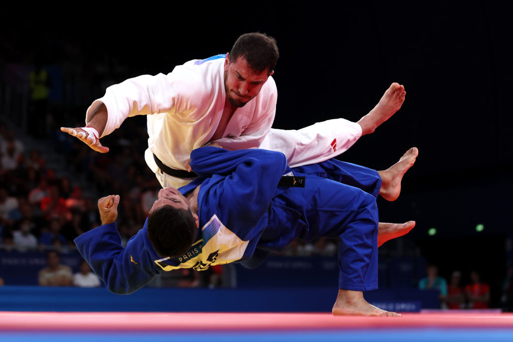 Judo practitioners, one in a white gi and the other in a blue gi, compete in a match. The athlete in the blue gi is mid-air, thrown by the one in the white gi