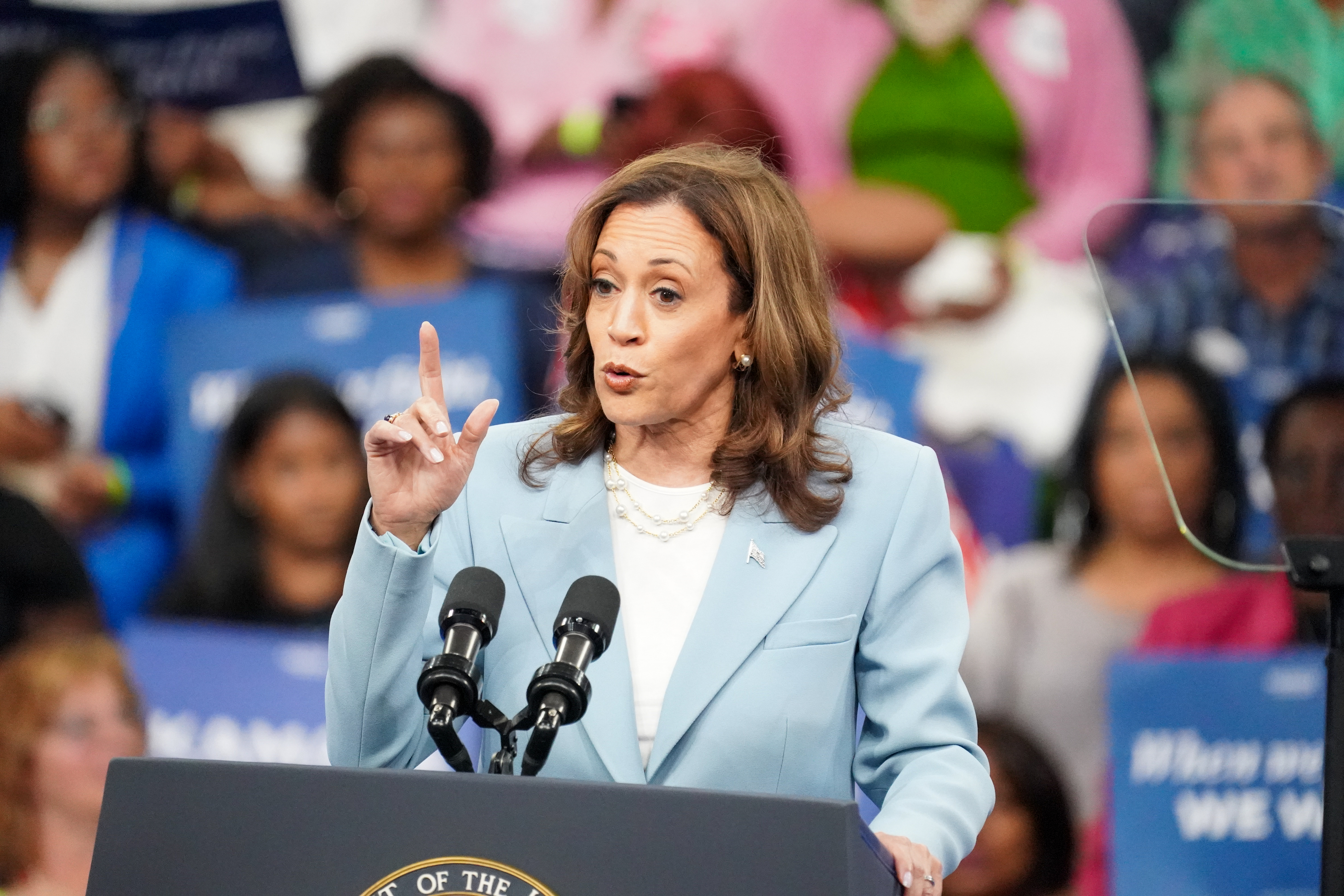 Kamala Harris speaks passionately at a podium during an event, gesturing with her right hand, with a diverse audience listening in the background