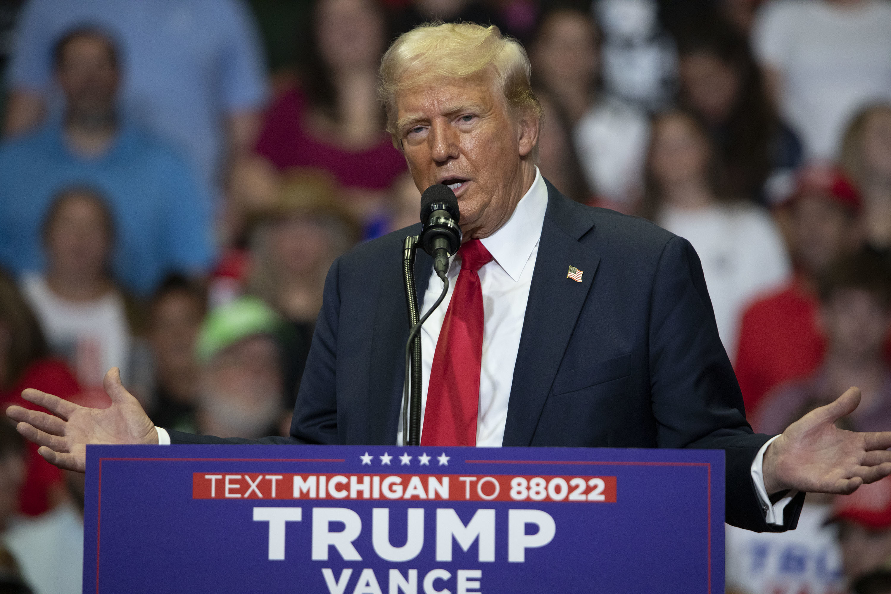 Donald Trump speaking at a rally in front of a crowd, standing behind a podium that reads &quot;Text MICHIGAN to 88022, TRUMP VANCE.&quot;