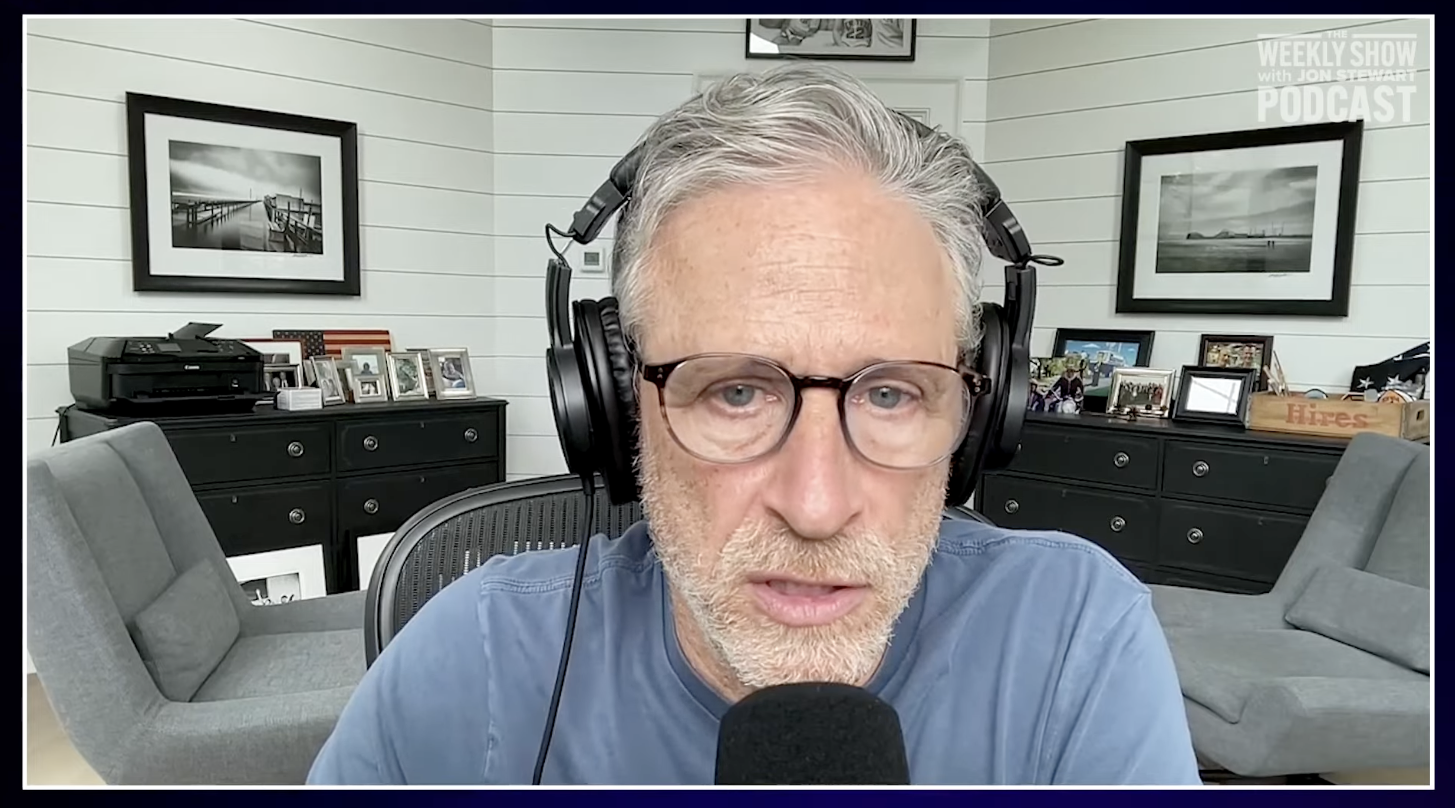 Jon Stewart sitting in a podcast setup with headphones and a microphone, in a room with framed photographs on the wall in the background