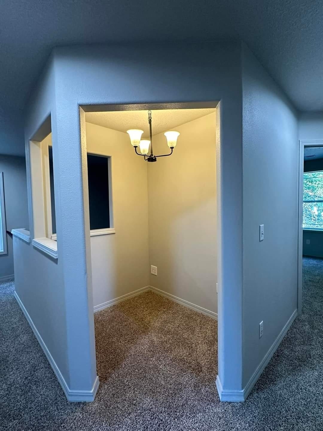 A small nook in a home interior with a hanging ceiling light and carpeted floor. The nook is next to a hallway with a window-like opening on the left wall