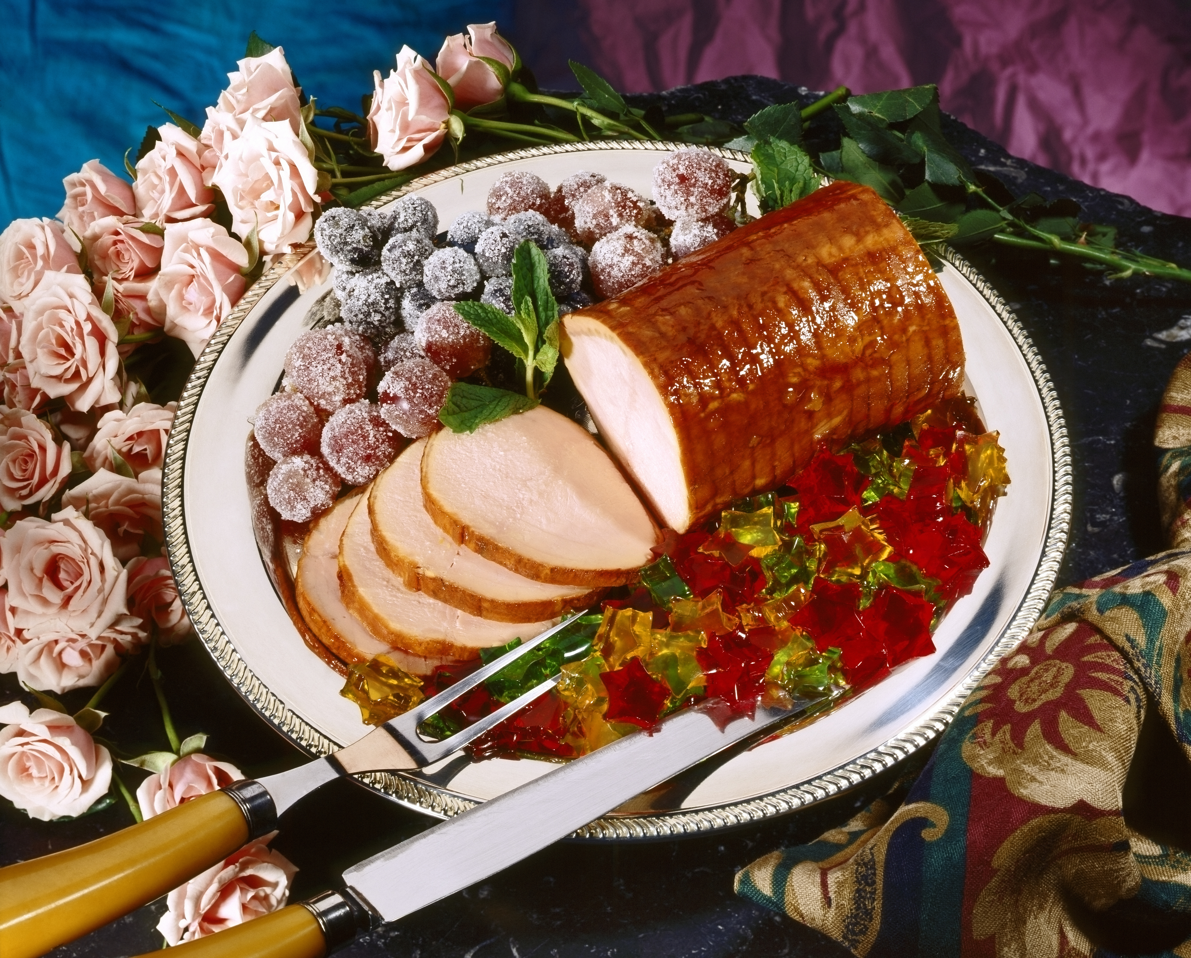 A neatly prepared platter with glazed ham slices, sugared grapes, colorful jellied candies, and pink roses for decoration. A fork and knife are on the platter