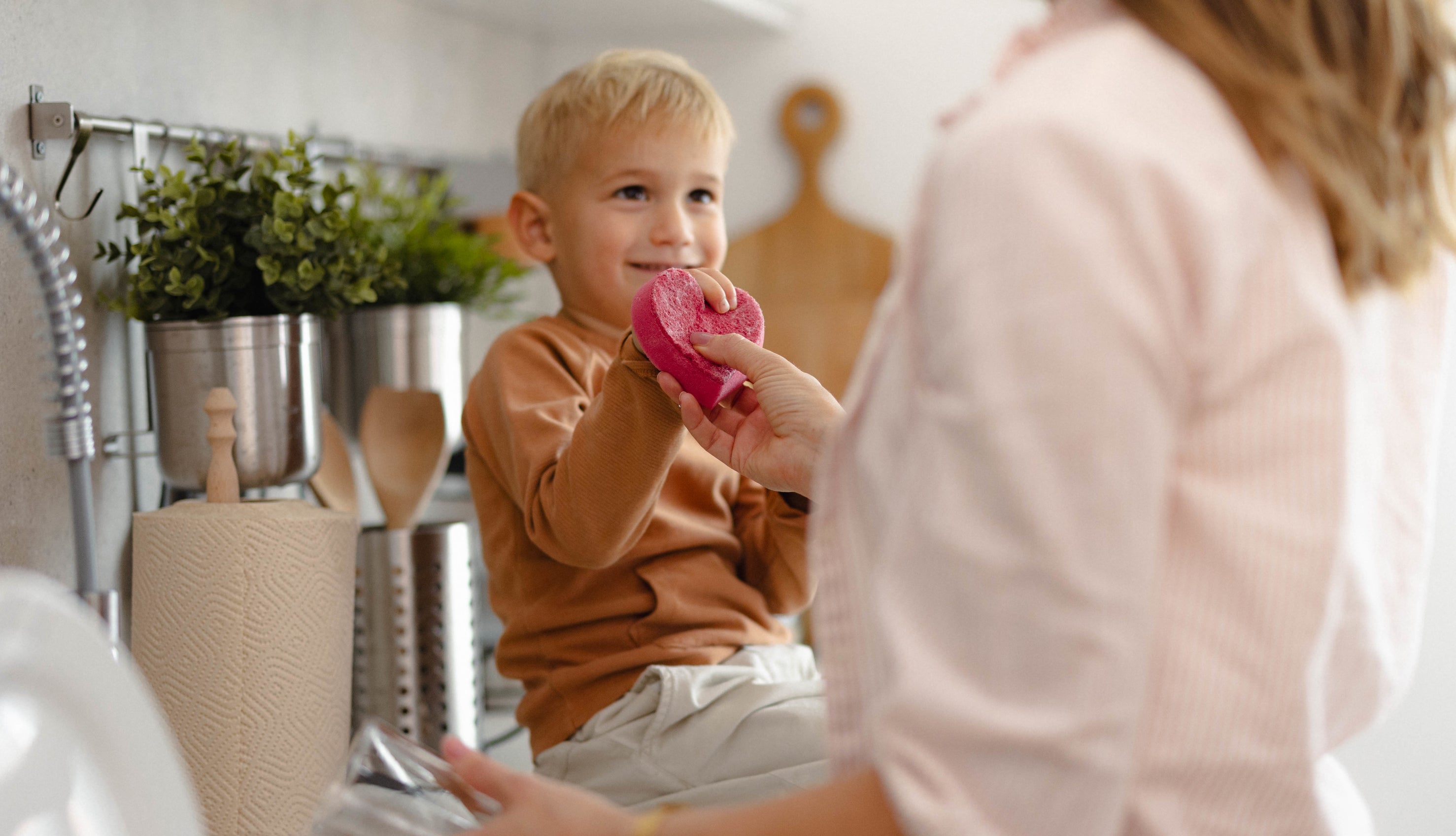 A woman helps a young blond boy holding a pink donut in a kitchen. The boy is sitting on the countertop near the sink