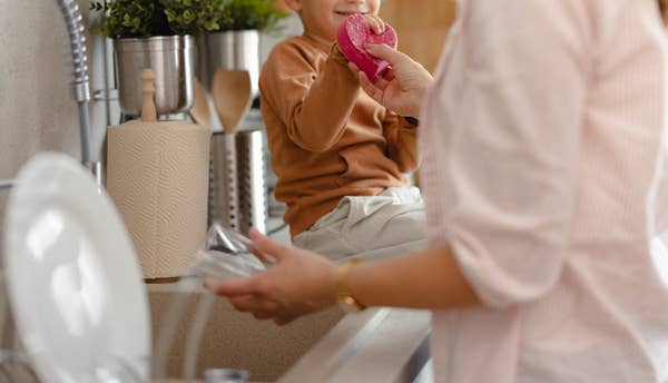 A woman helps a young blond boy holding a pink donut in a kitchen. The boy is sitting on the countertop near the sink