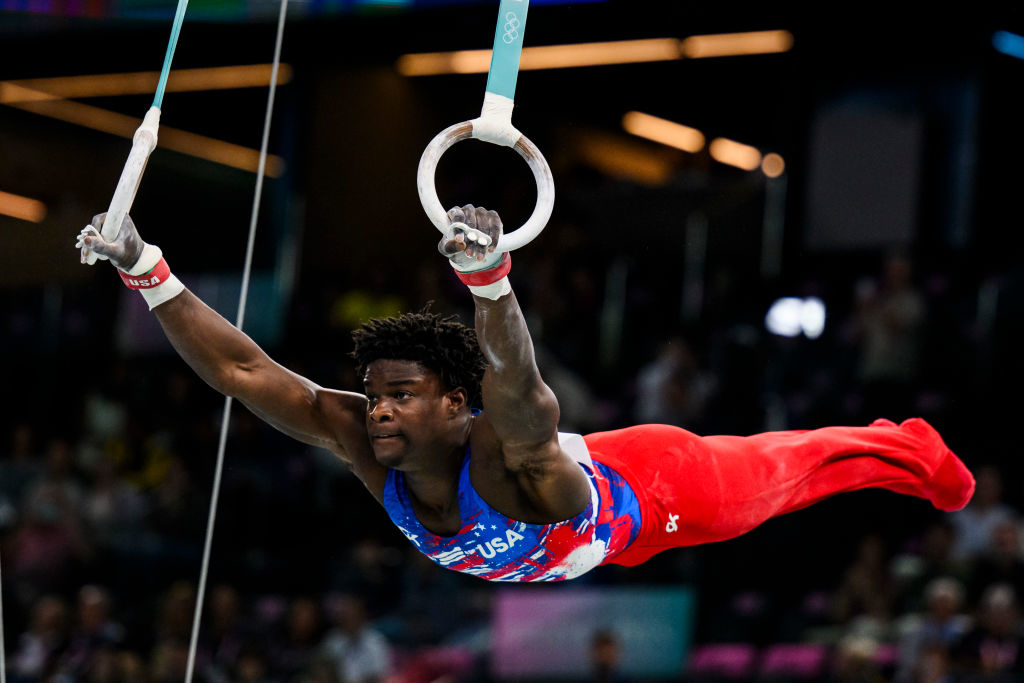 Gymnast Donnell Whittenburg performs a routine on the rings, wearing a sleeveless USA jersey and red pants, focusing intently during competition