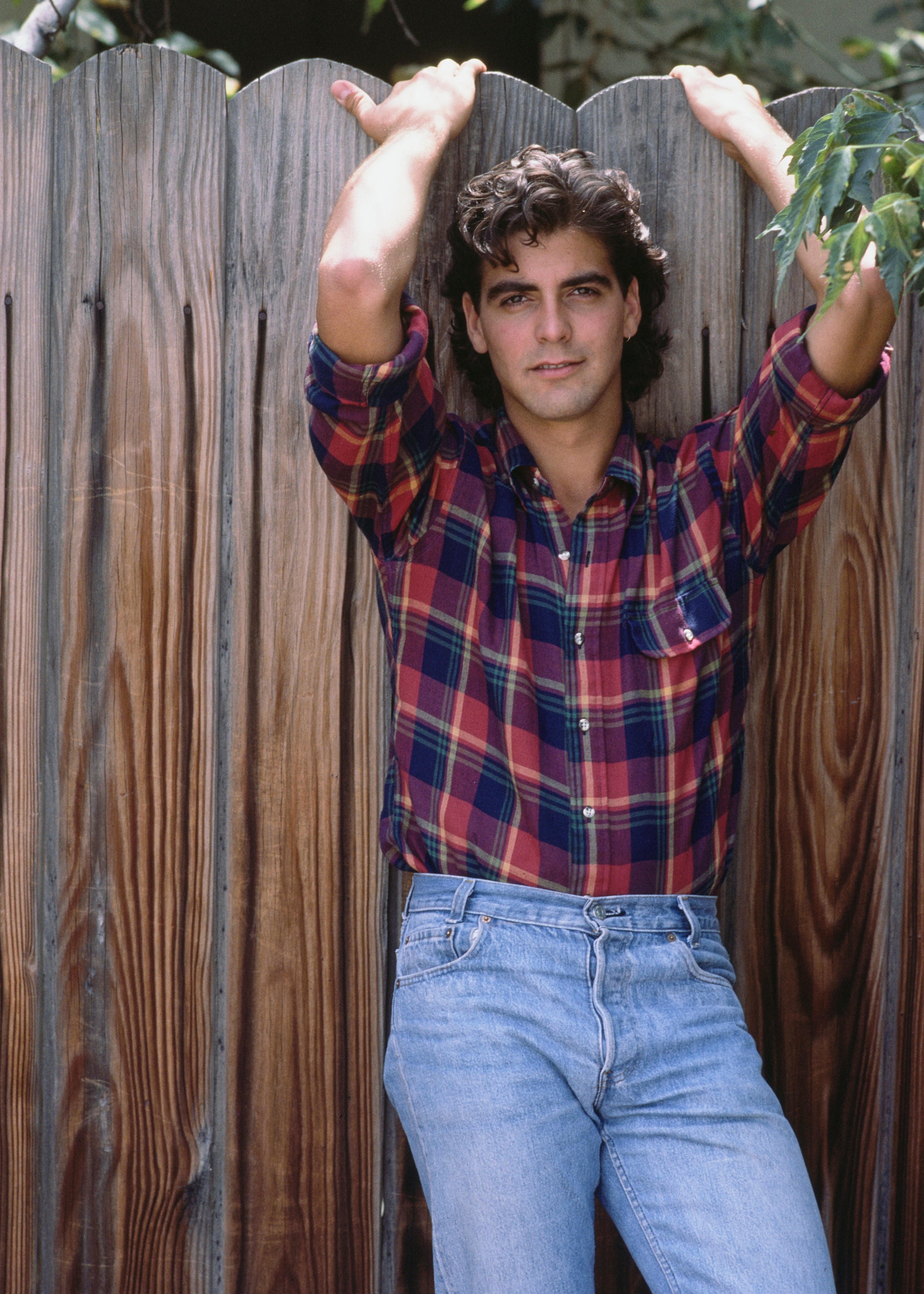 George Clooney leaning against a wooden fence, wearing a plaid shirt and jeans