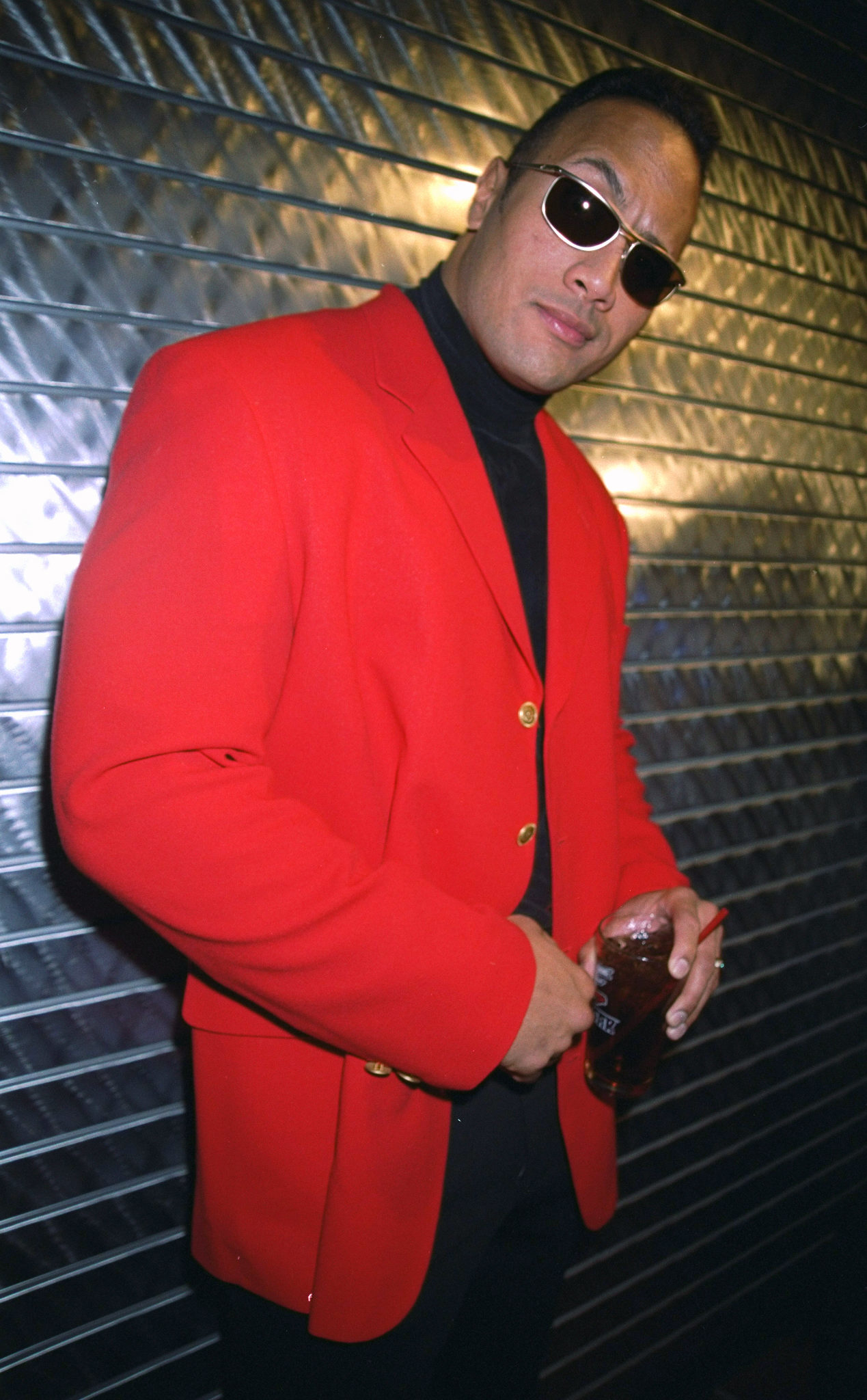 Dwayne Johnson is seen in a red blazer over a black turtleneck, wearing sunglasses and holding a drink