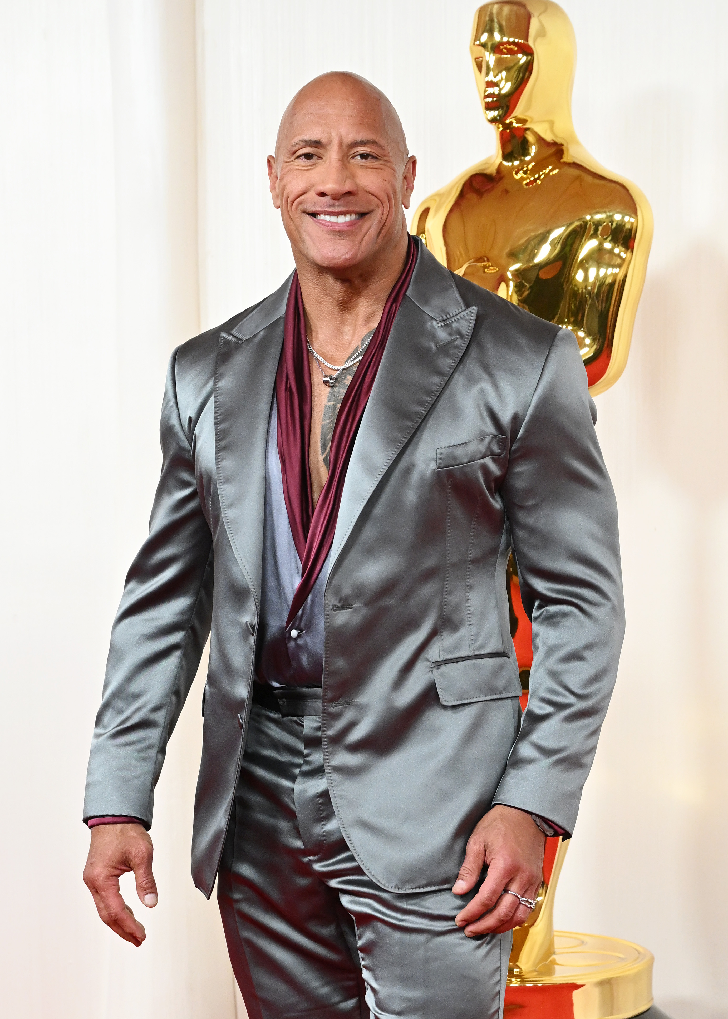 Dwayne Johnson at an event, dressed in a satin suit with a deep V-neck shirt, standing in front of an Oscar statue