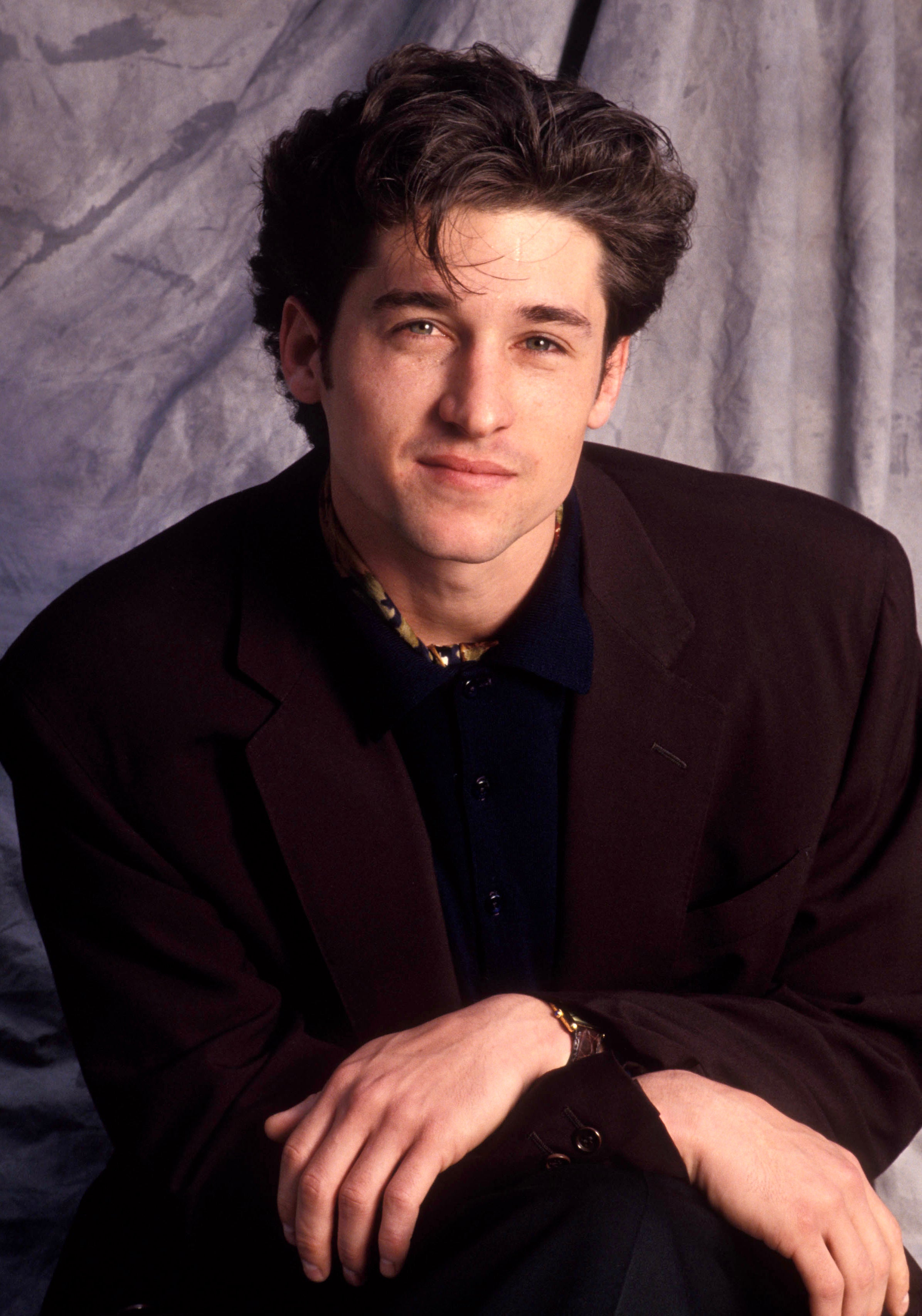 Patrick Dempsey poses in a studio with a relaxed smile, wearing a dark blazer over a collared shirt, against a draped fabric background