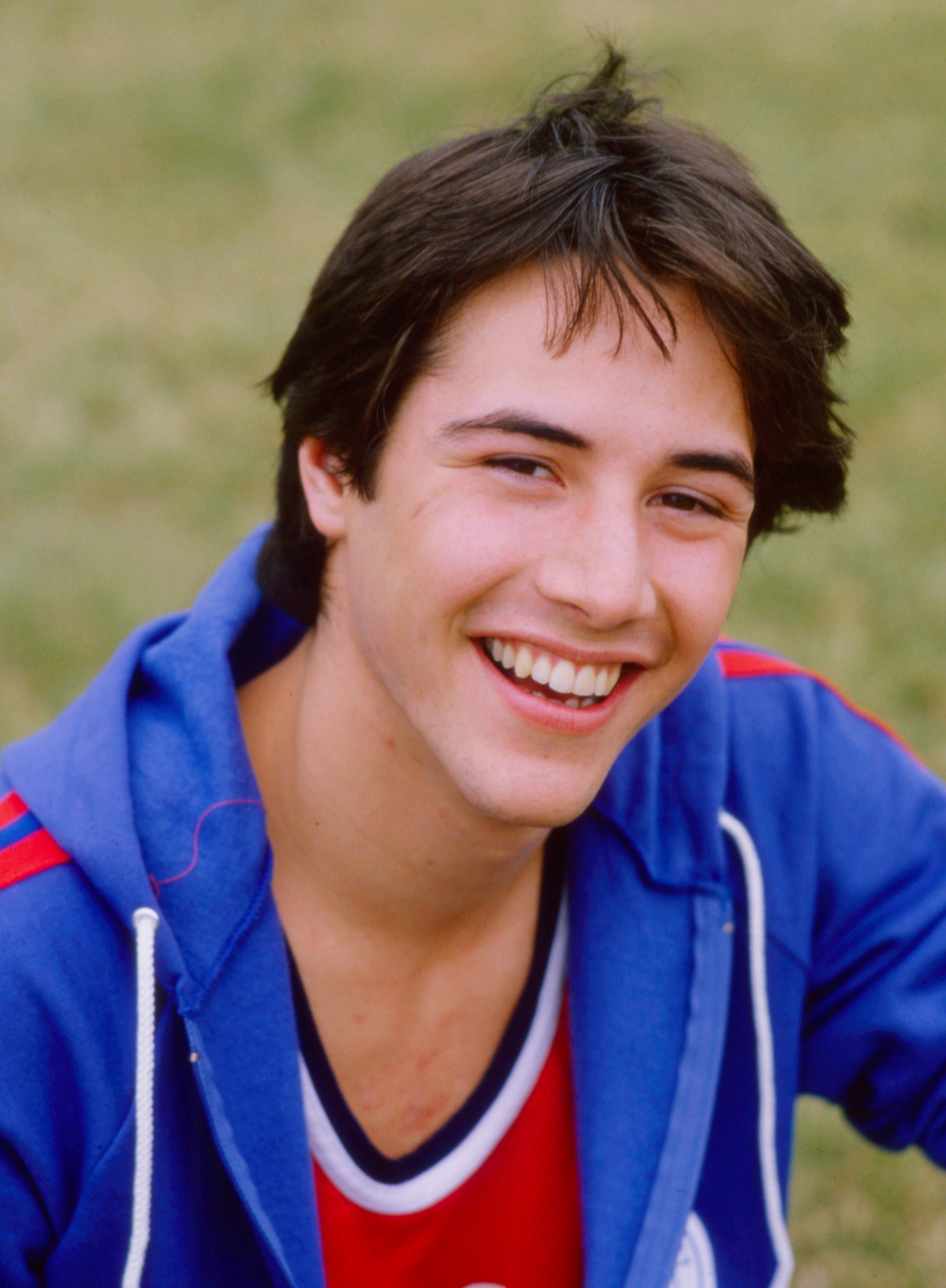 Keanu Reeves smiling, wearing a casual athletic jacket and top while sitting outdoors