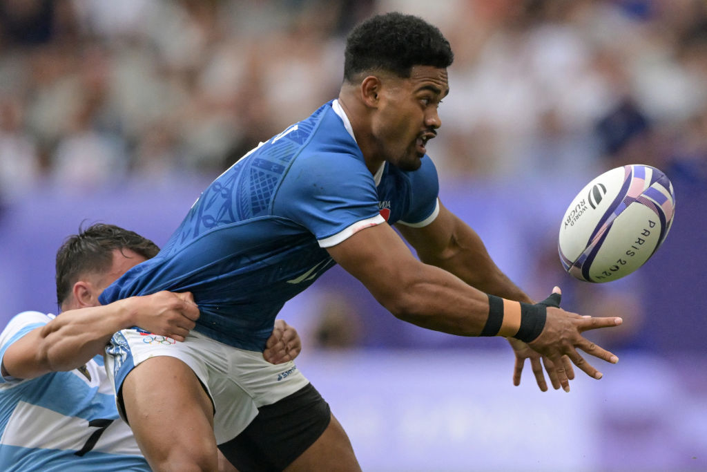 Rugby player drops the ball as an opposing team member tackles him during a match. The background shows an outdoor stadium packed with spectators
