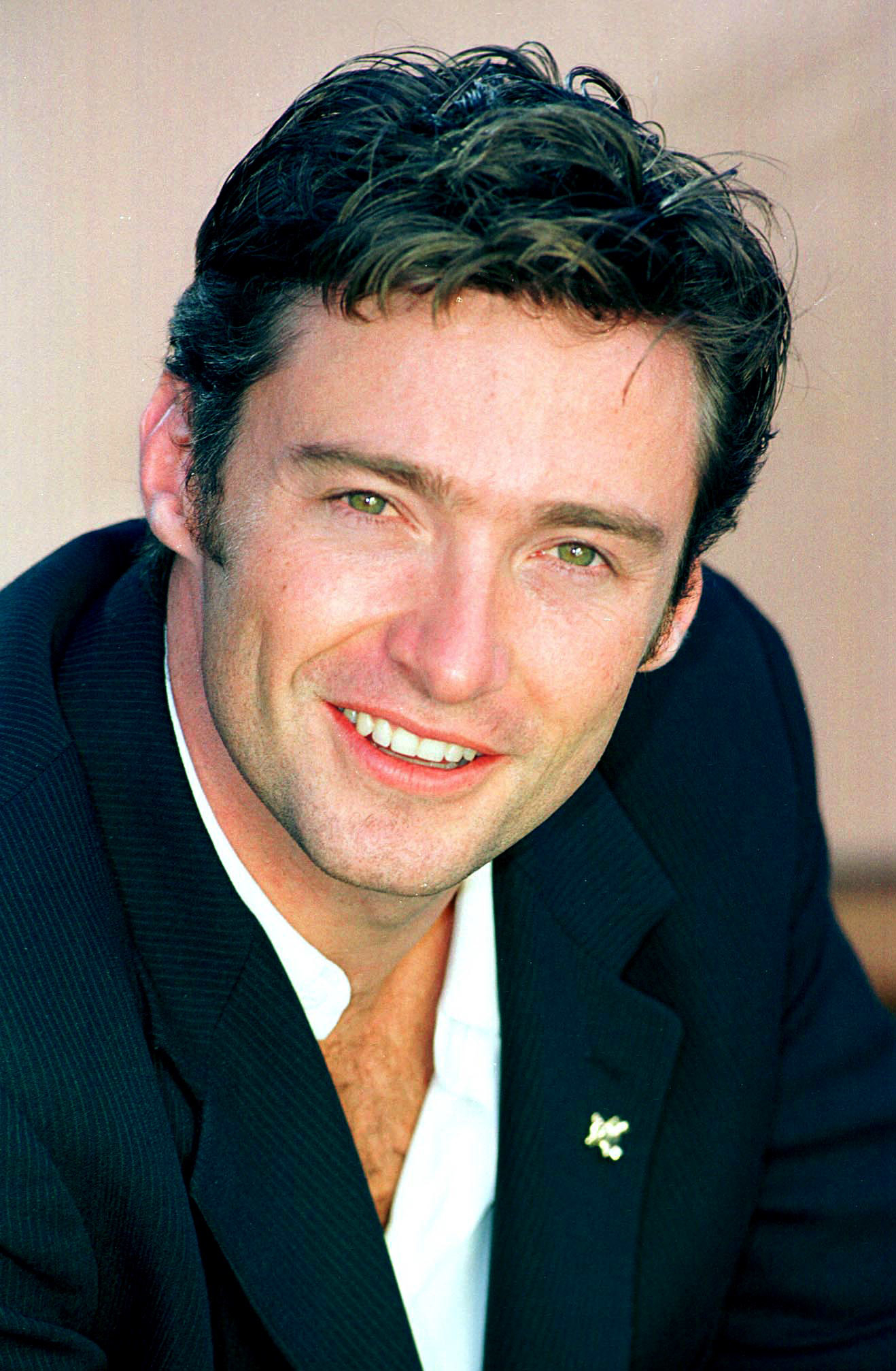 Hugh Jackman, in a smart-casual black blazer and white shirt, smiles warmly at the camera