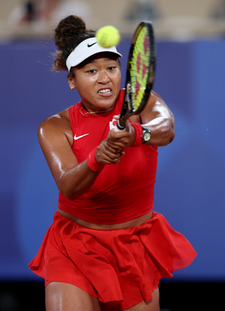 Naomi Osaka playing tennis, focused on hitting the ball with her racket. She is wearing a red athletic outfit with a headband