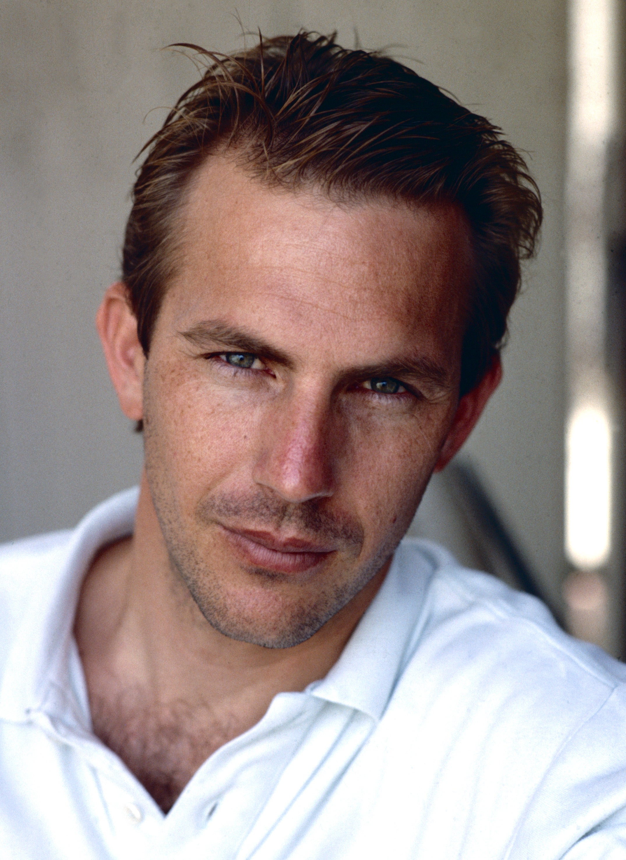 Kevin Costner, wearing a white collared shirt, looks directly into the camera with a slight smile